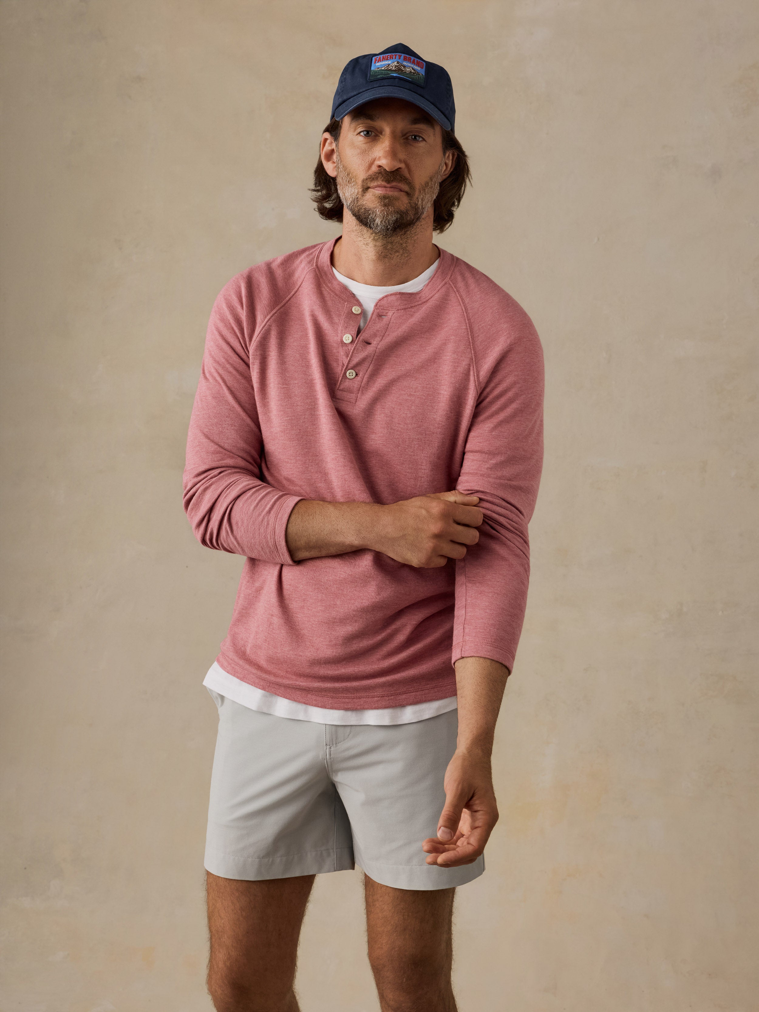 A man wearing a dusty rose long-sleeve henley shirt, light gray shorts, a navy blue baseball cap, and a white undershirt, standing against a textured beige wall.