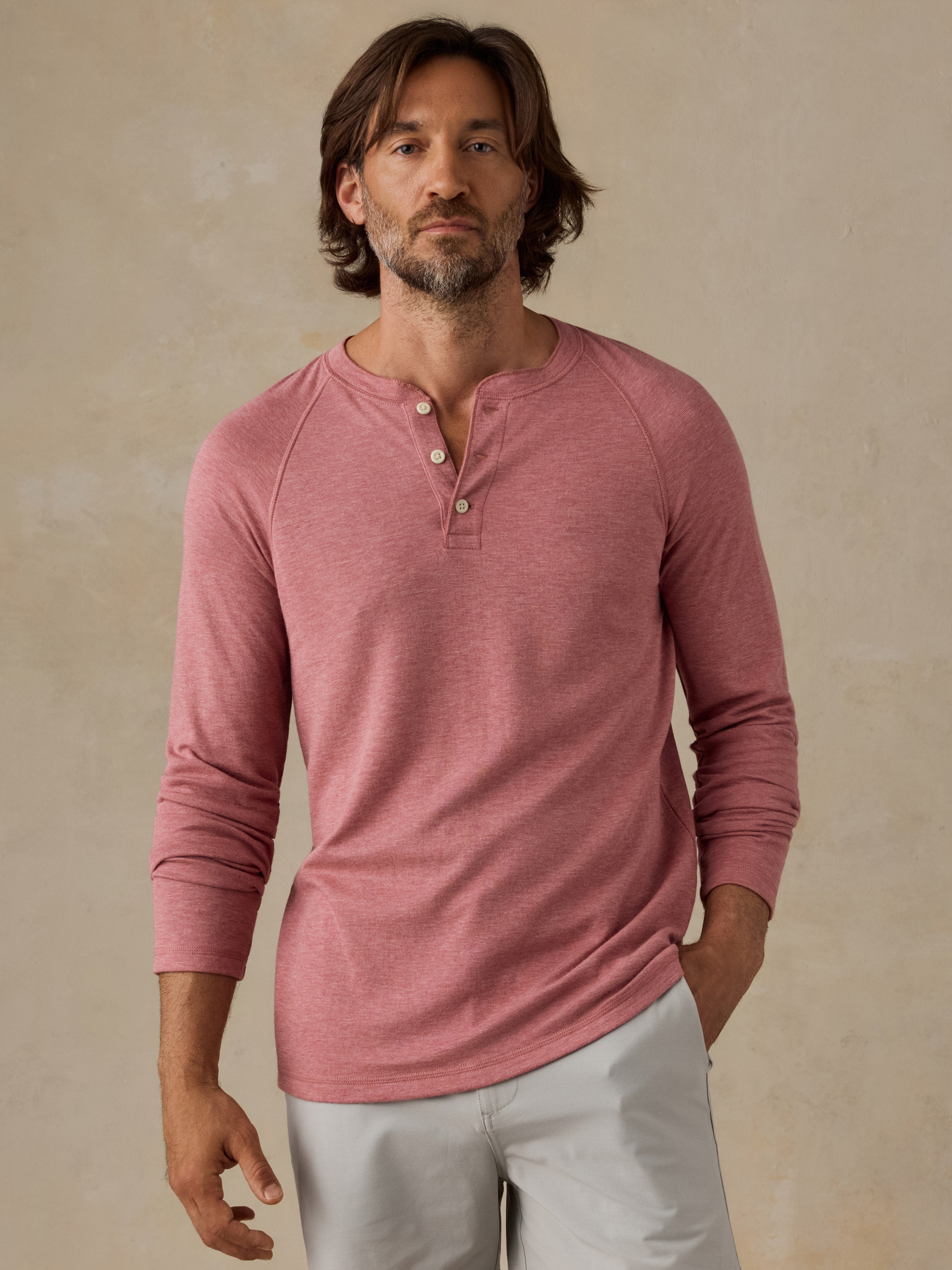 A man wearing a dusty rose pink long-sleeve henley shirt with grey shorts, standing against a textured beige wall.