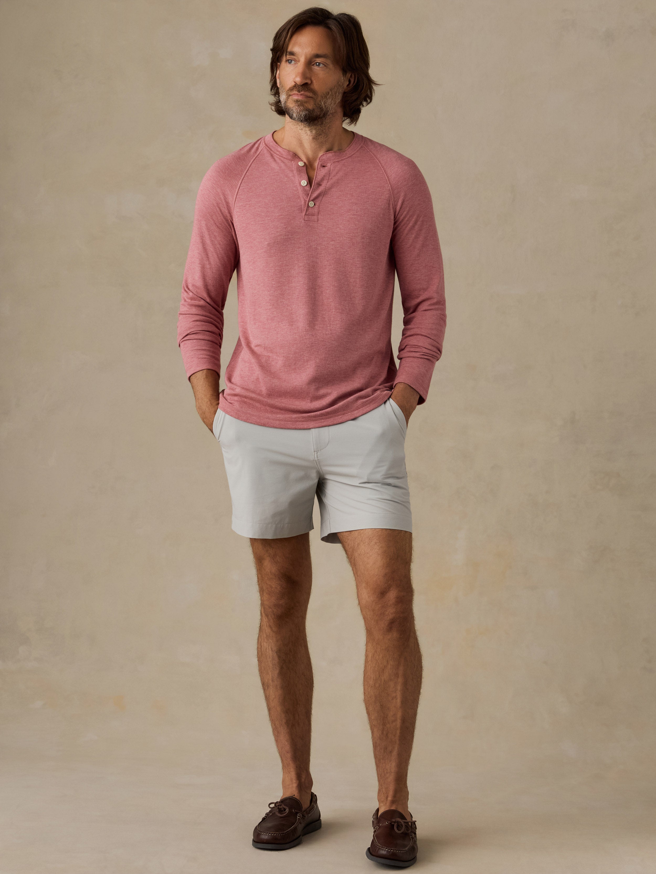 A man wearing a dusty rose long-sleeve henley shirt, light gray shorts, and dark brown loafers, standing against a textured beige studio backdrop.