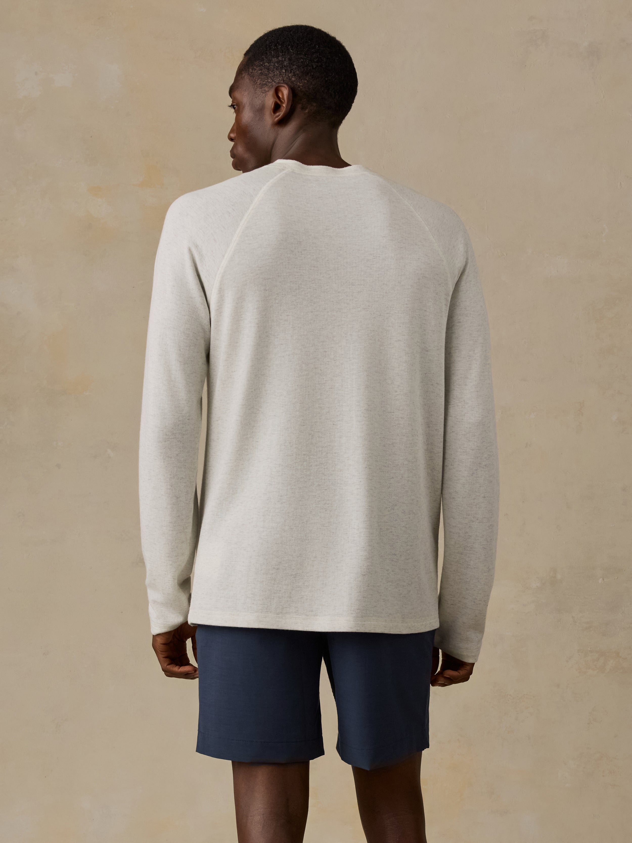 Man is wearing a cream colored long-sleeve henley shirt and navy shorts against a beige background