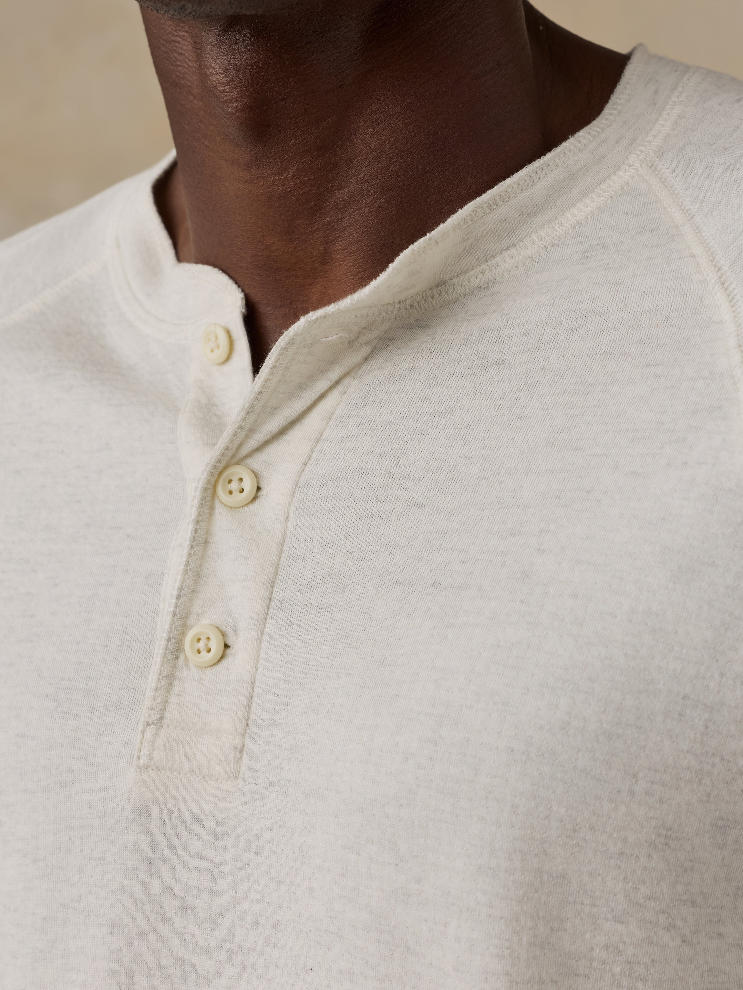 Man is wearing a cream colored long-sleeve henley shirt against a beige background