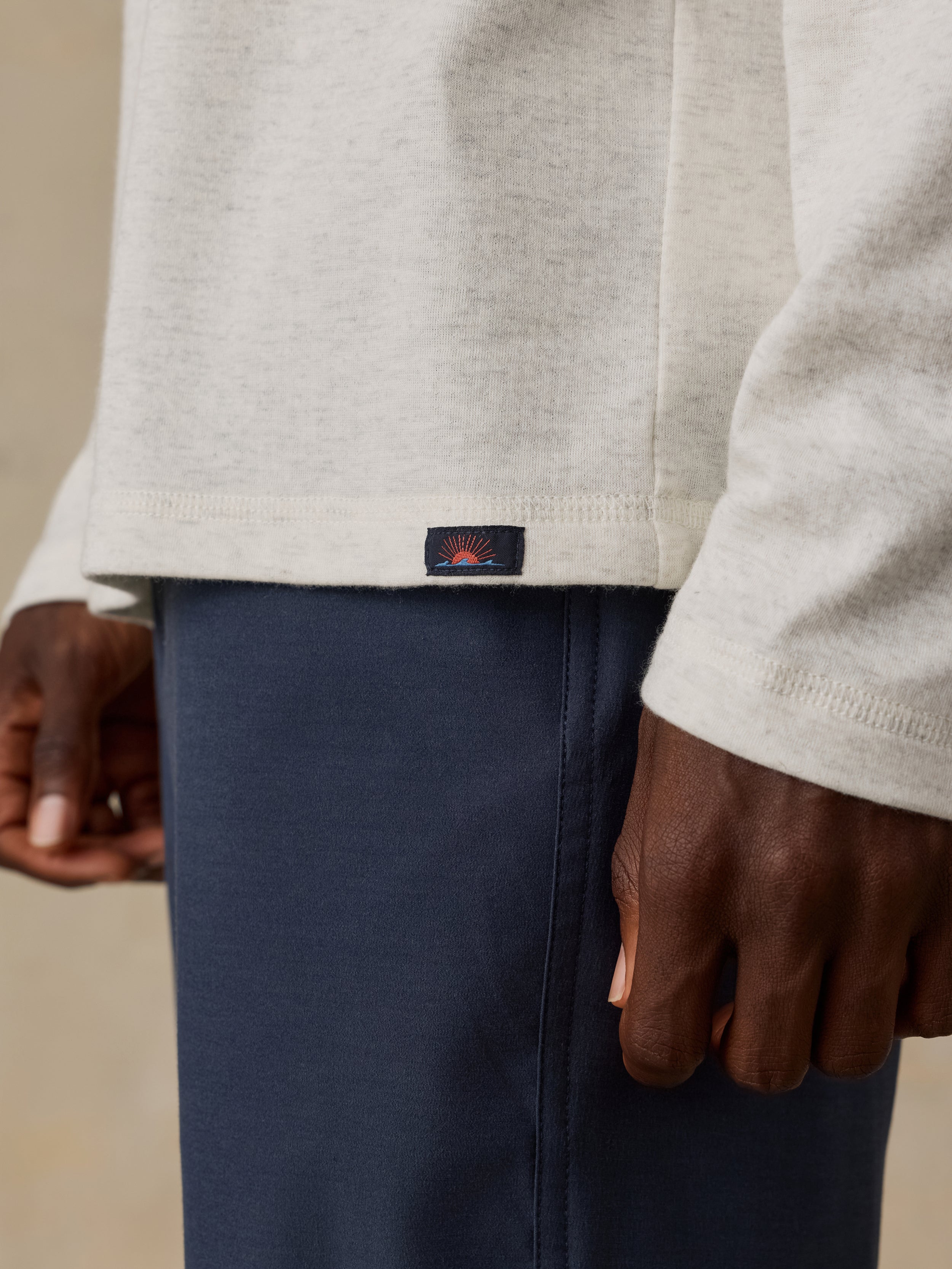 Close-up of a person wearing a cream colored long-sleeve top and navy shorts, with a small Faherty brand label near the hem of the shirt.