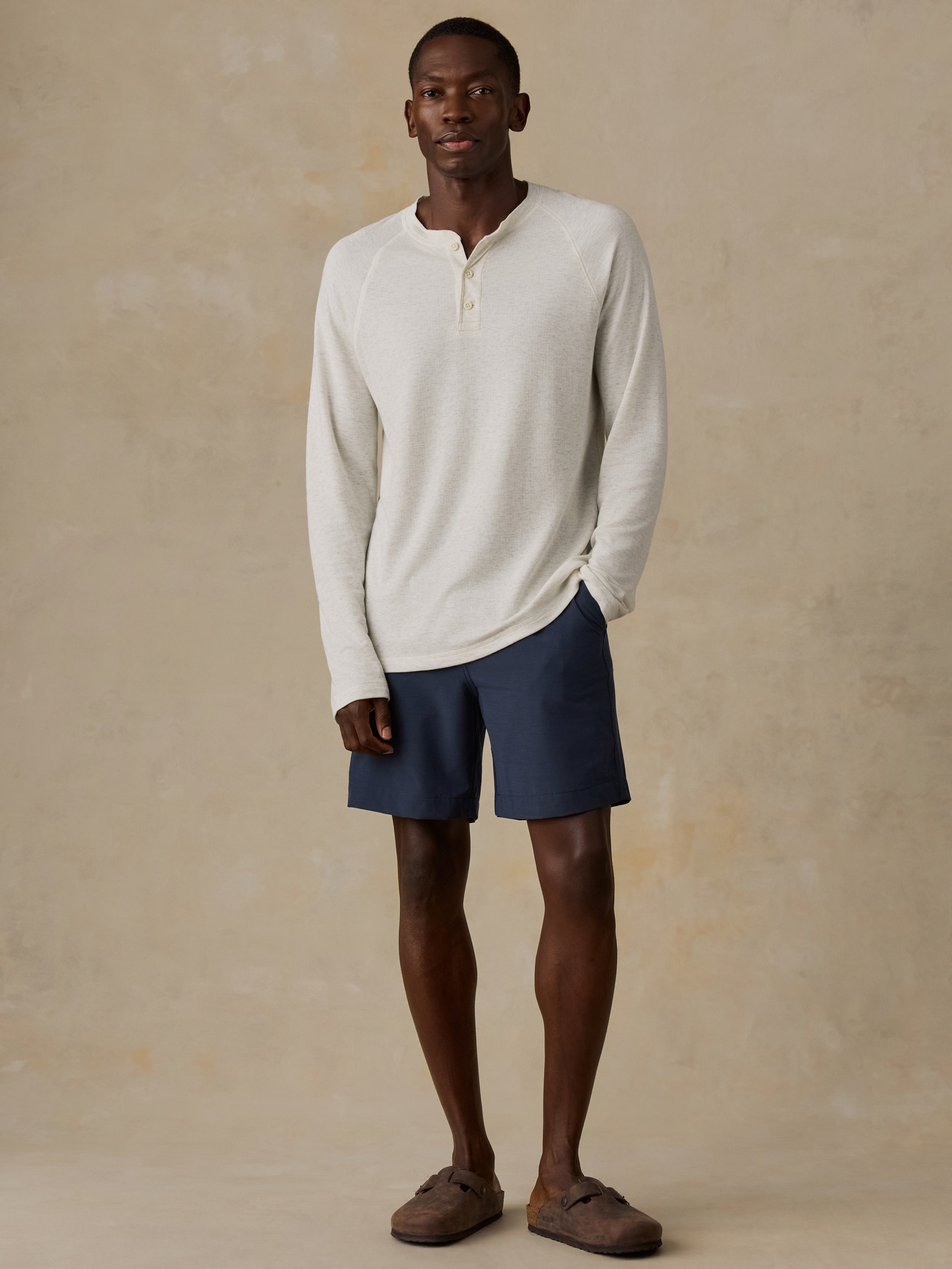 Man is wearing a cream colored long-sleeve henley shirt and navy shorts against a beige background