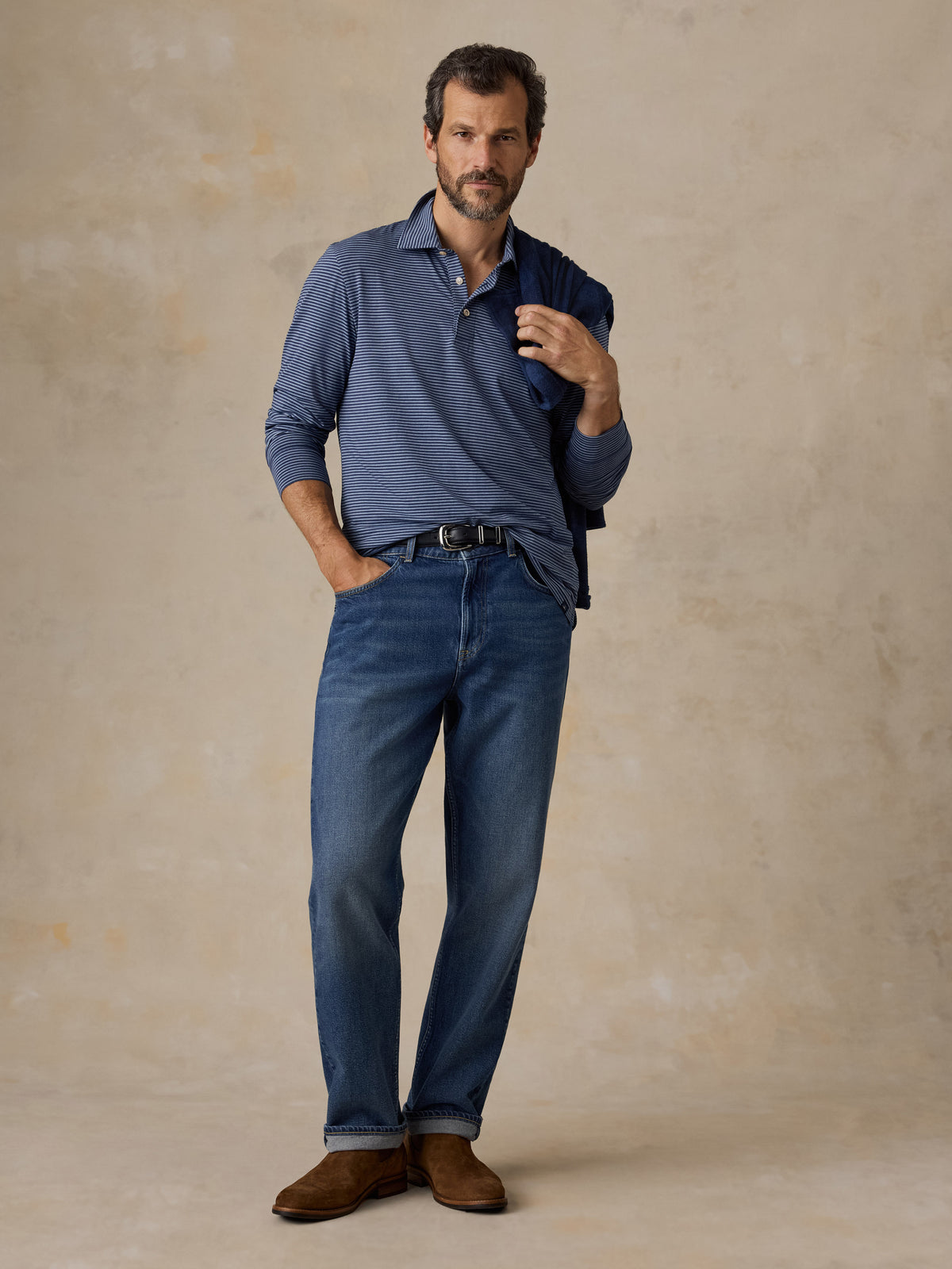 A man wearing a blue striped polo shirt, blue jeans, and suede boots, image 2 of 5