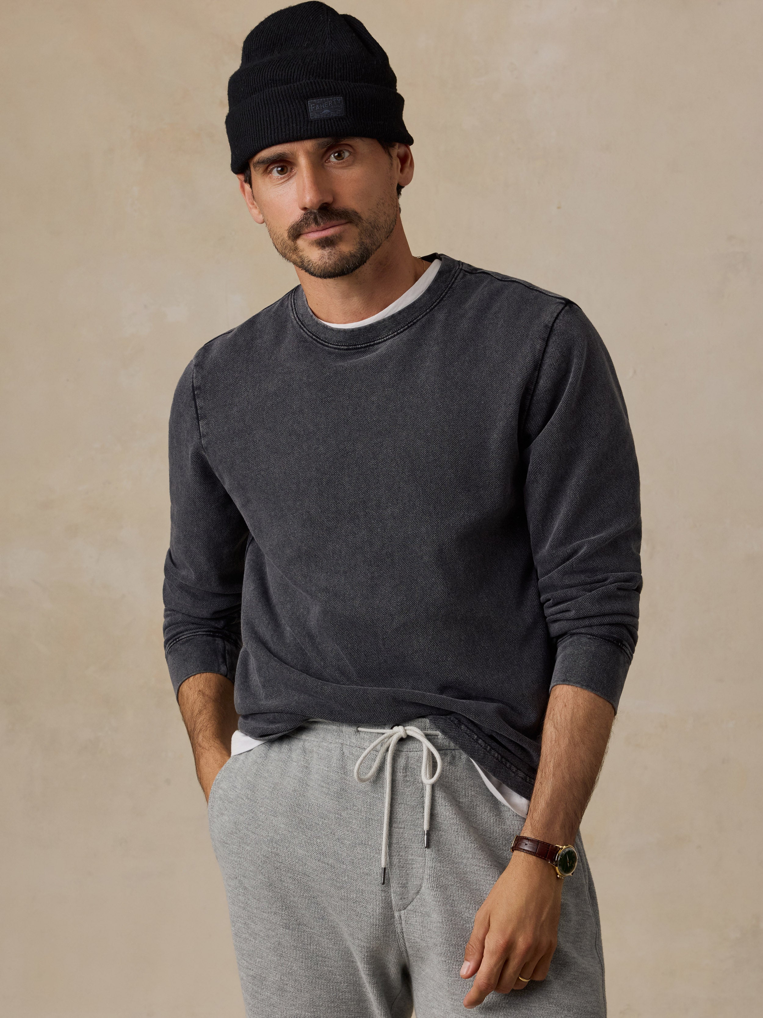A man wearing a dark charcoal gray long-sleeve crewneck sweatshirt, light gray sweatpants, a black knit beanie, and a brown leather watch, standing against a textured beige studio background.