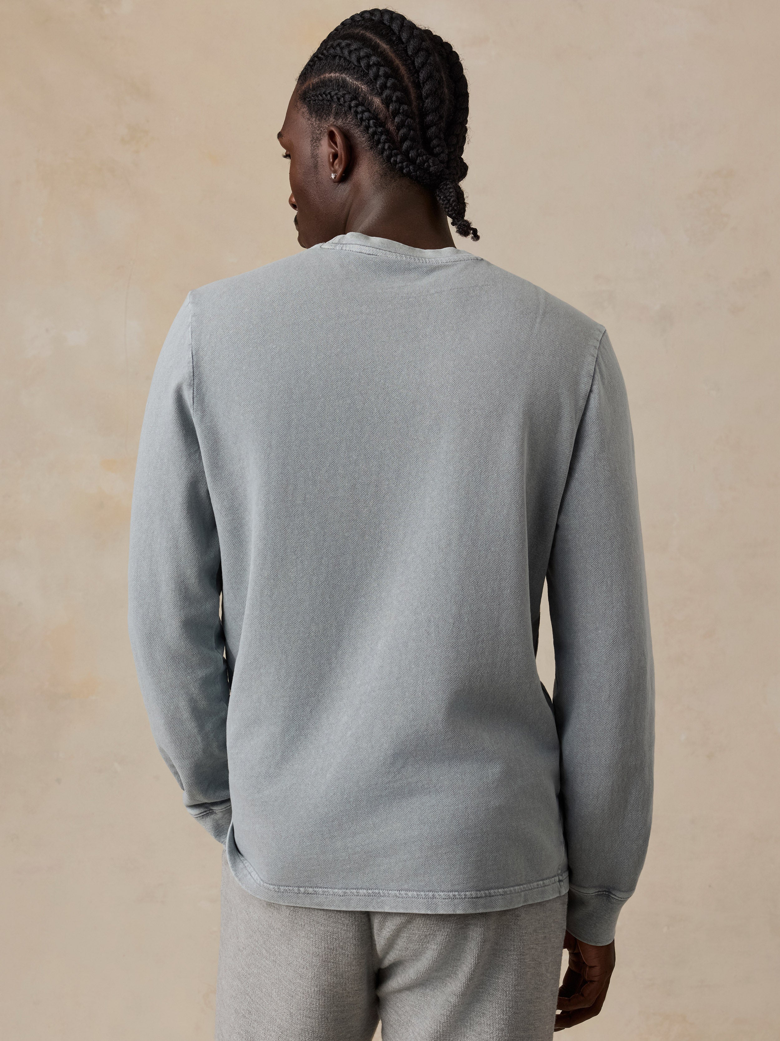 A man viewed from behind wearing a light gray ribbed long-sleeve crewneck sweatshirt and matching gray sweatpants, standing against a textured beige background.