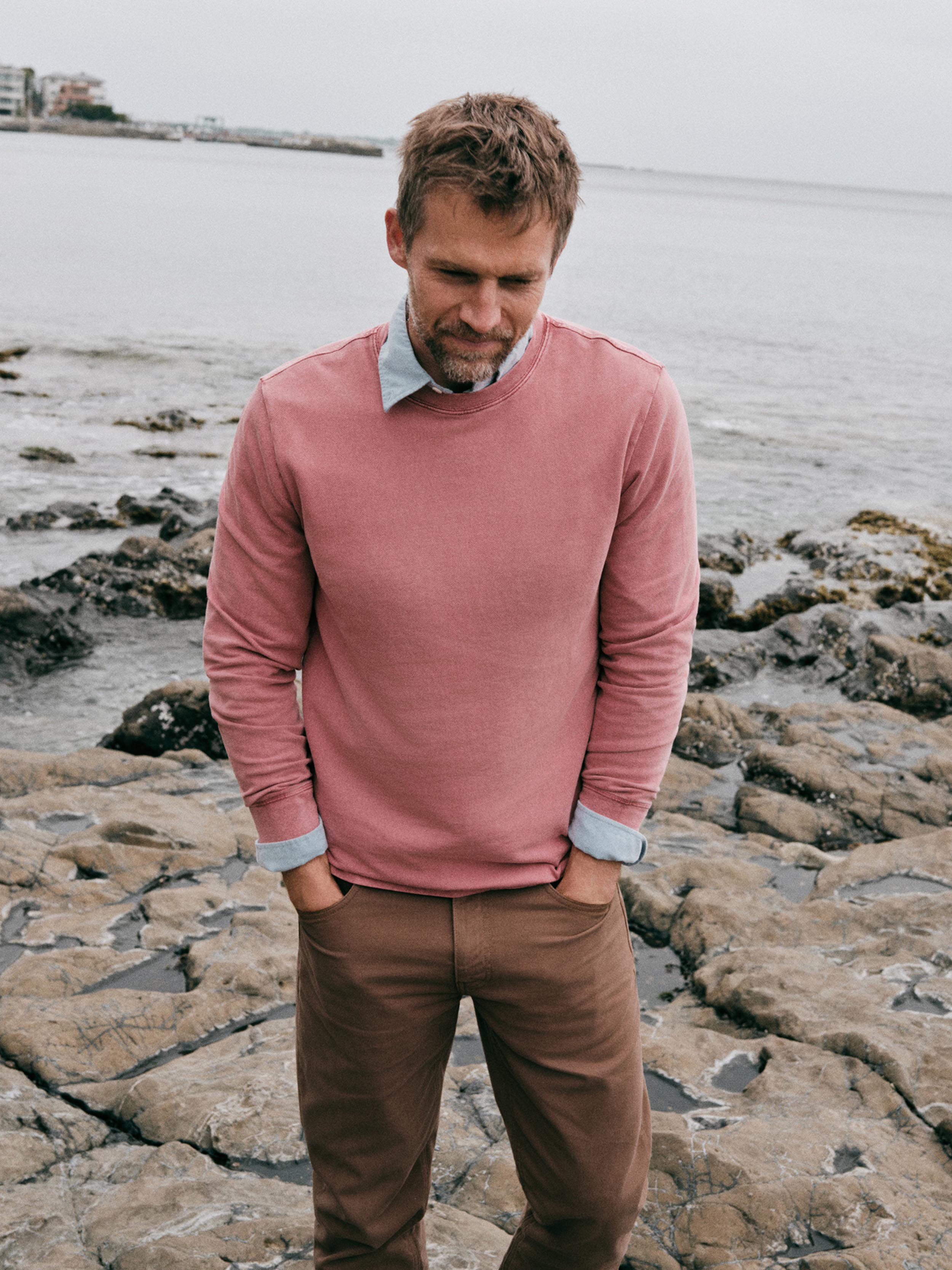 A man standing on rocky shoreline wearing a dusty rose long-sleeve crewneck sweater over a light blue collared shirt, paired with brown trousers.