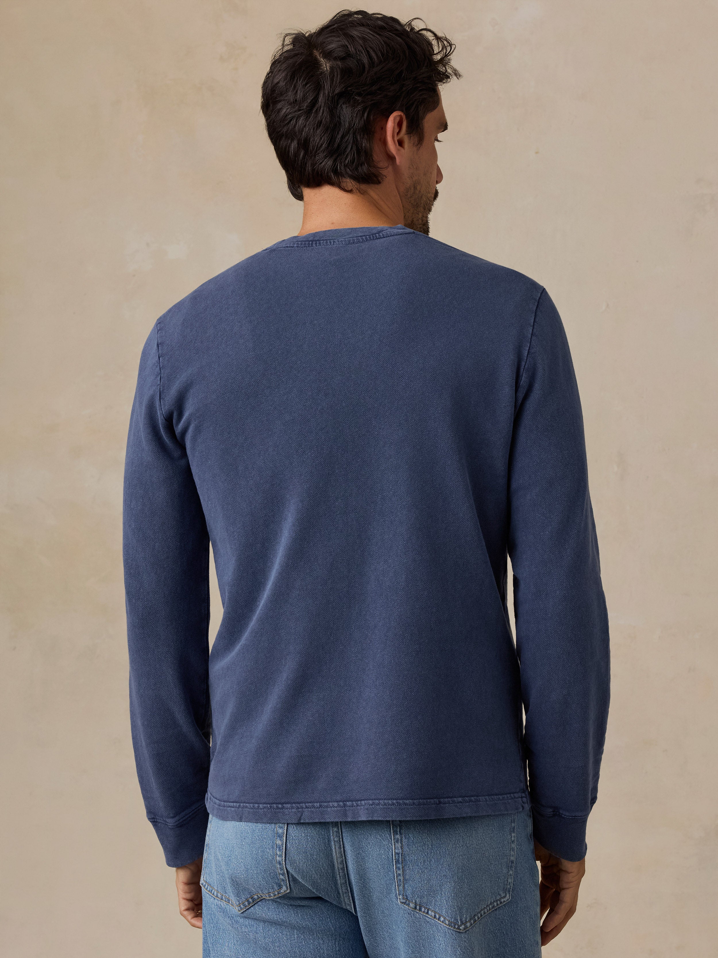 A man viewed from behind wearing a navy blue long-sleeve ribbed knit shirt and light blue jeans, standing against a textured beige studio wall.