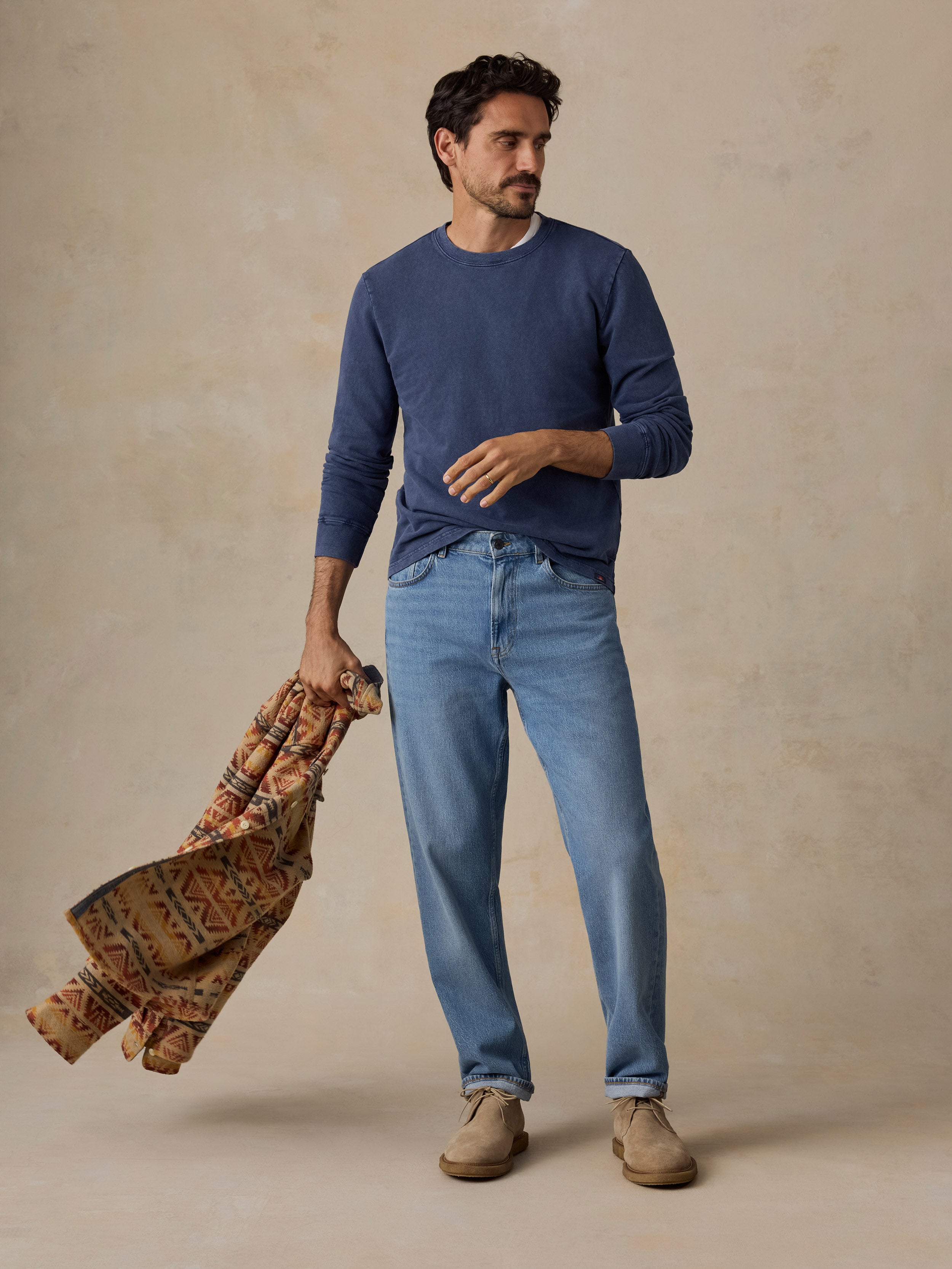 A man stands against a textured beige studio backdrop wearing a navy long-sleeve crewneck sweater, light blue jeans, and tan shoes, while holding a patterned brown and orange scarf.