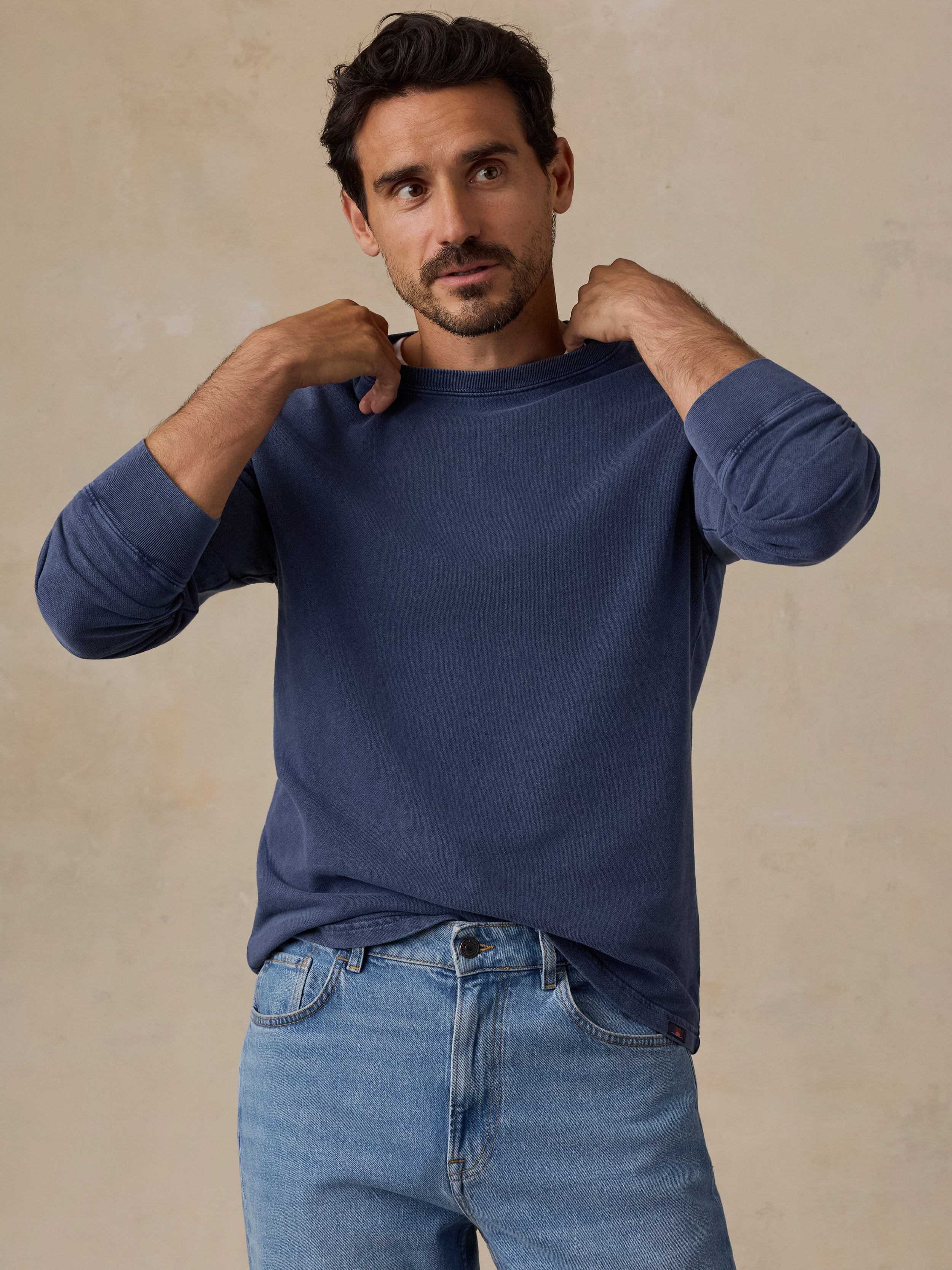 A man wearing a navy blue long-sleeve shirt and light blue jeans, posing with his hands on his shoulders against a textured beige background.