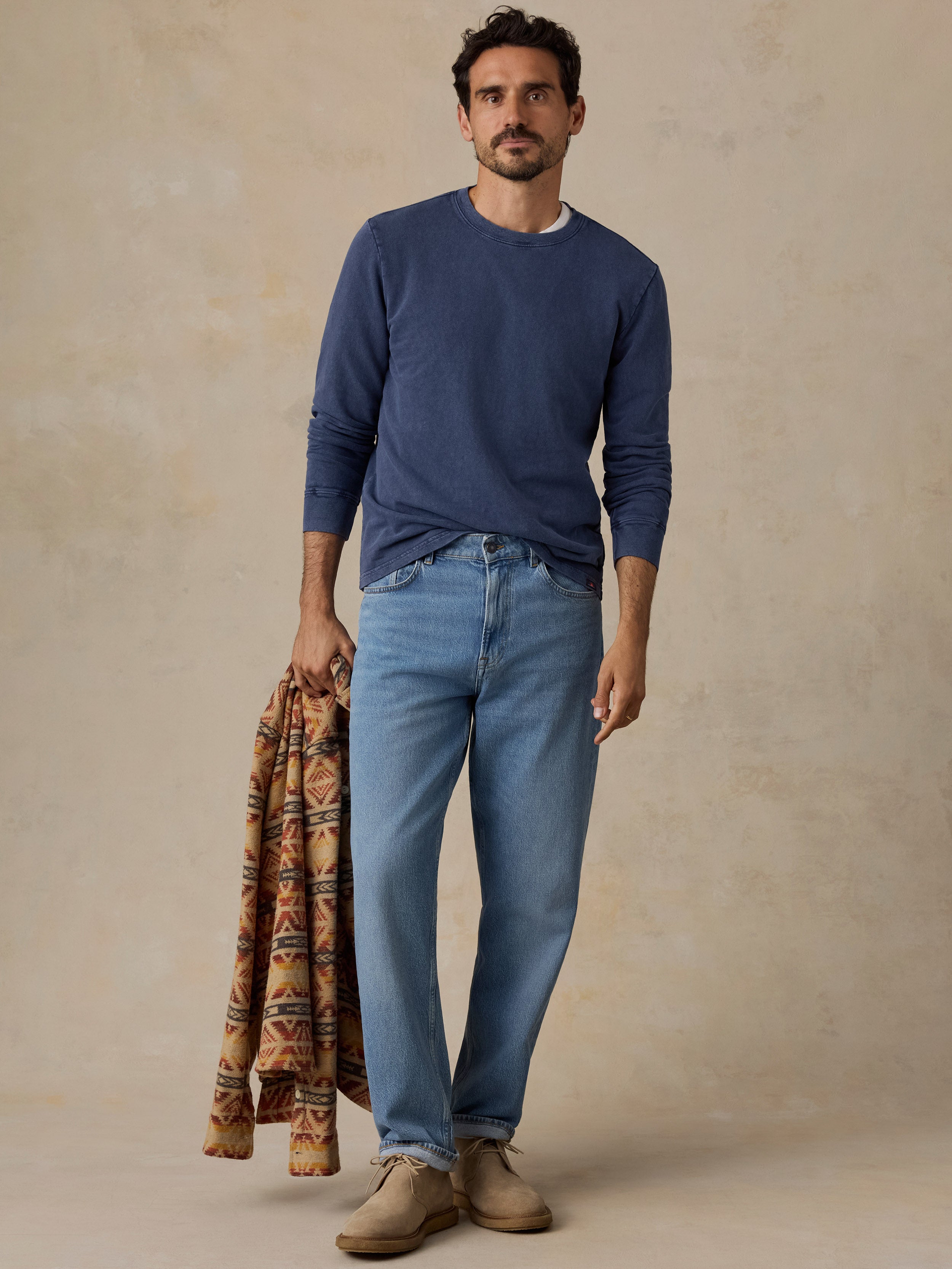 A man stands against a textured beige studio backdrop wearing a navy long-sleeve crewneck sweater, light blue jeans, and tan shoes, while holding a patterned brown and orange knit sweater draped over one arm.