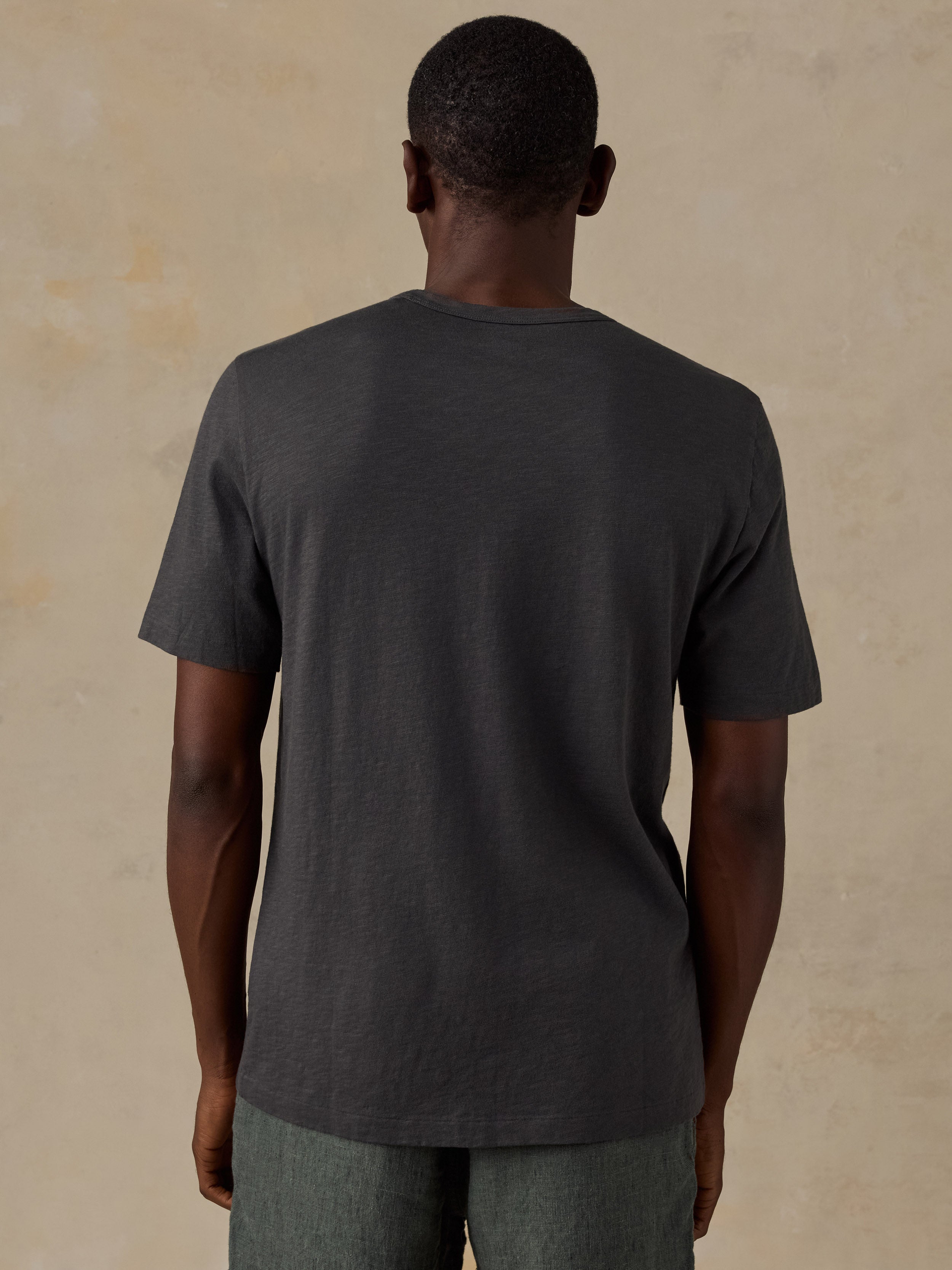 A man viewed from behind wearing a dark charcoal gray short-sleeve crewneck t-shirt and gray pants, standing against a textured beige background.