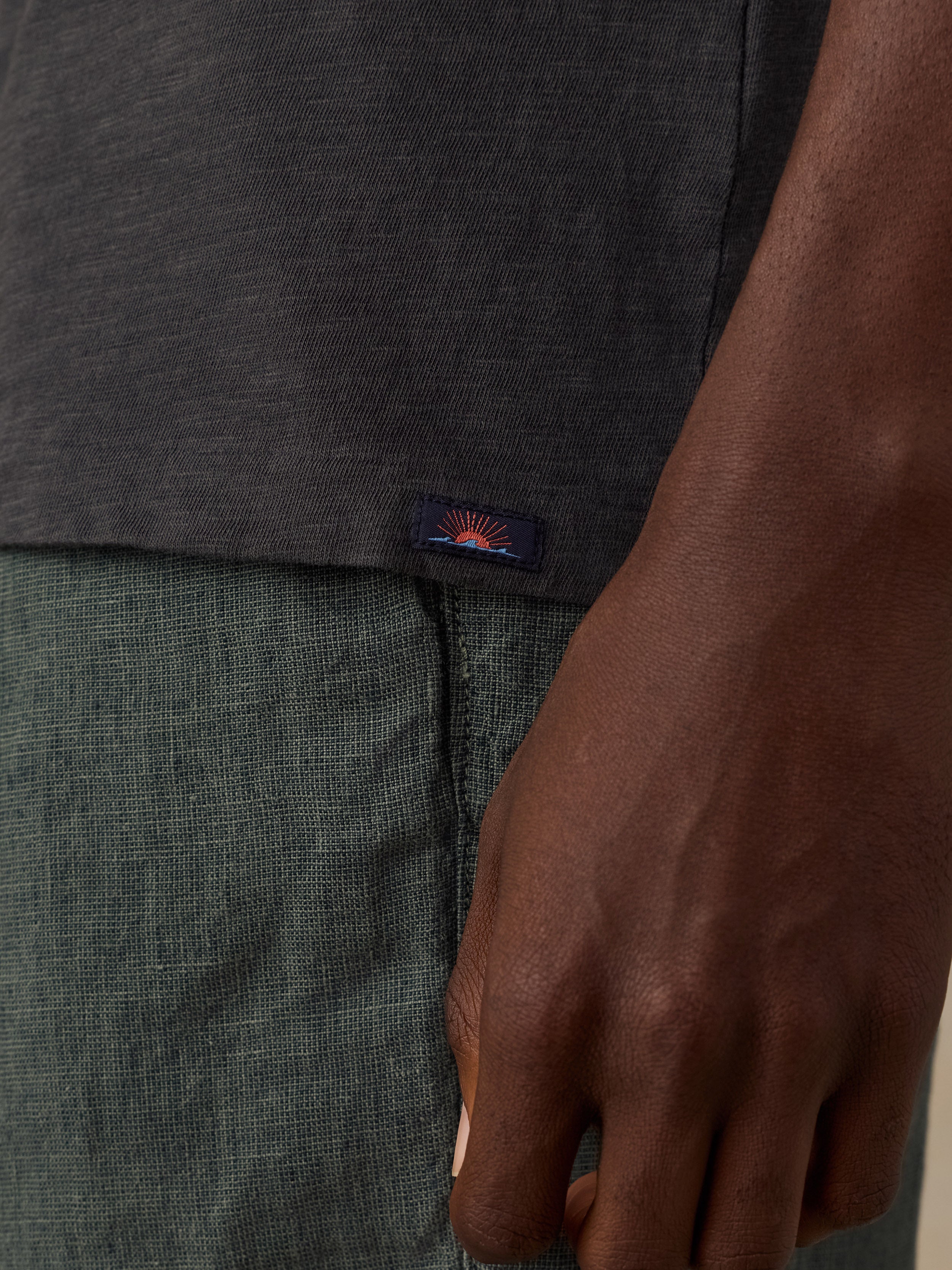 Close-up of a person wearing a dark charcoal gray heathered t-shirt and textured green-gray shorts, with a small embroidered Faherty brand sun and waves patch on the shirt hem.
