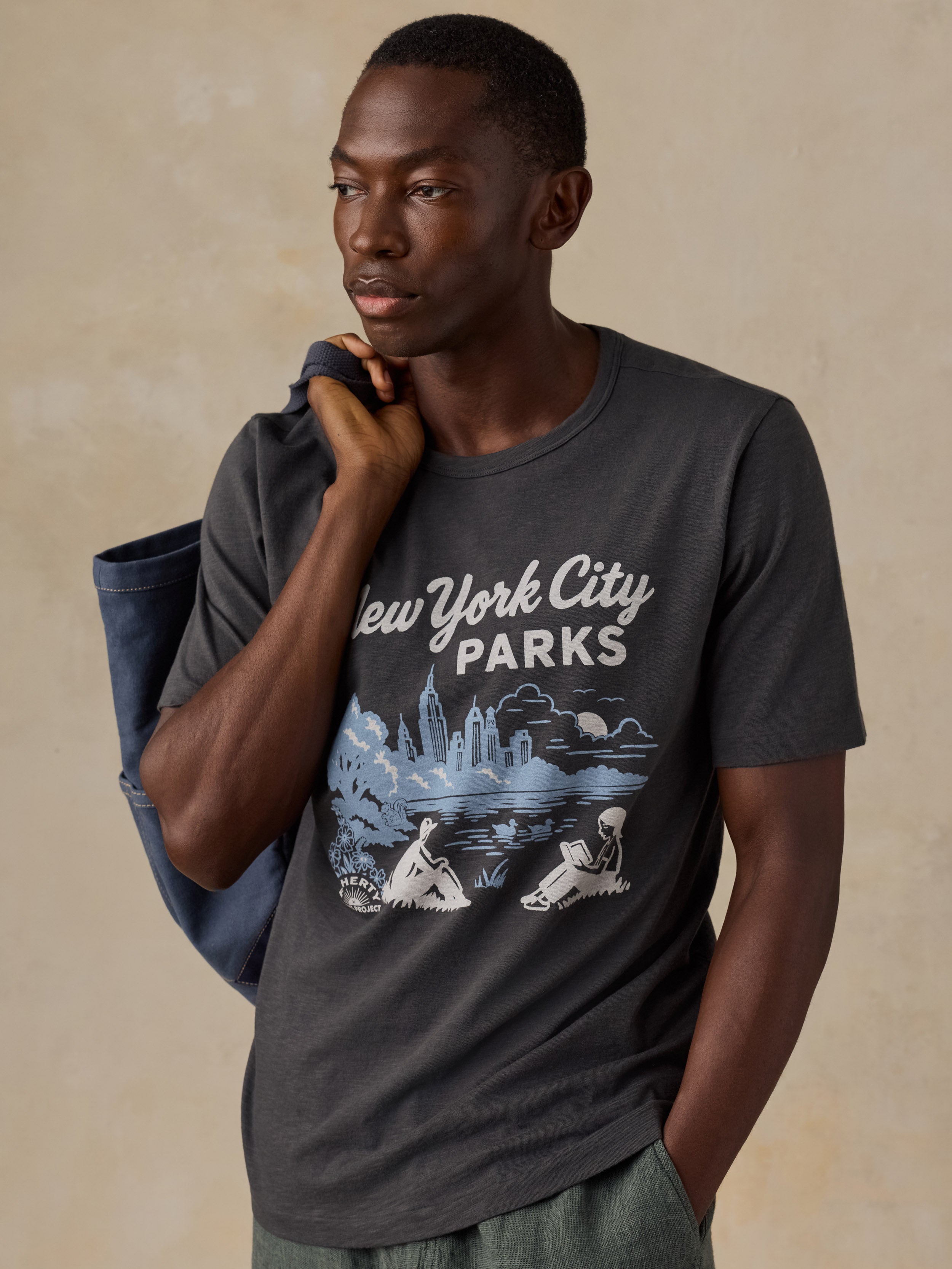 A man wearing a dark gray crewneck t-shirt with a 'New York City Parks' graphic print on the front, posed against a textured beige background.