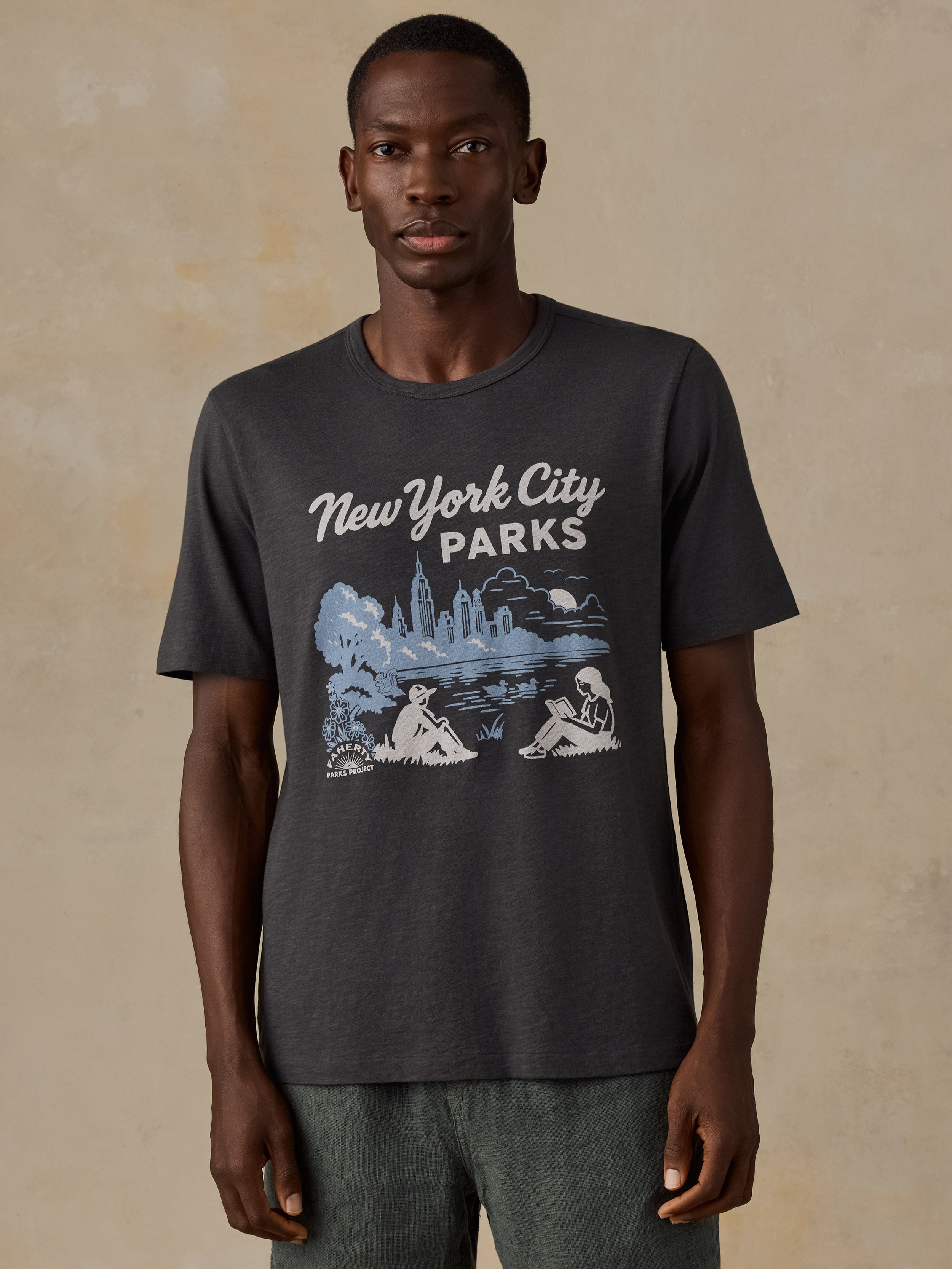 A man wearing a dark charcoal gray t-shirt with a 'New York City Parks' graphic print in white and light blue, standing against a textured beige background.