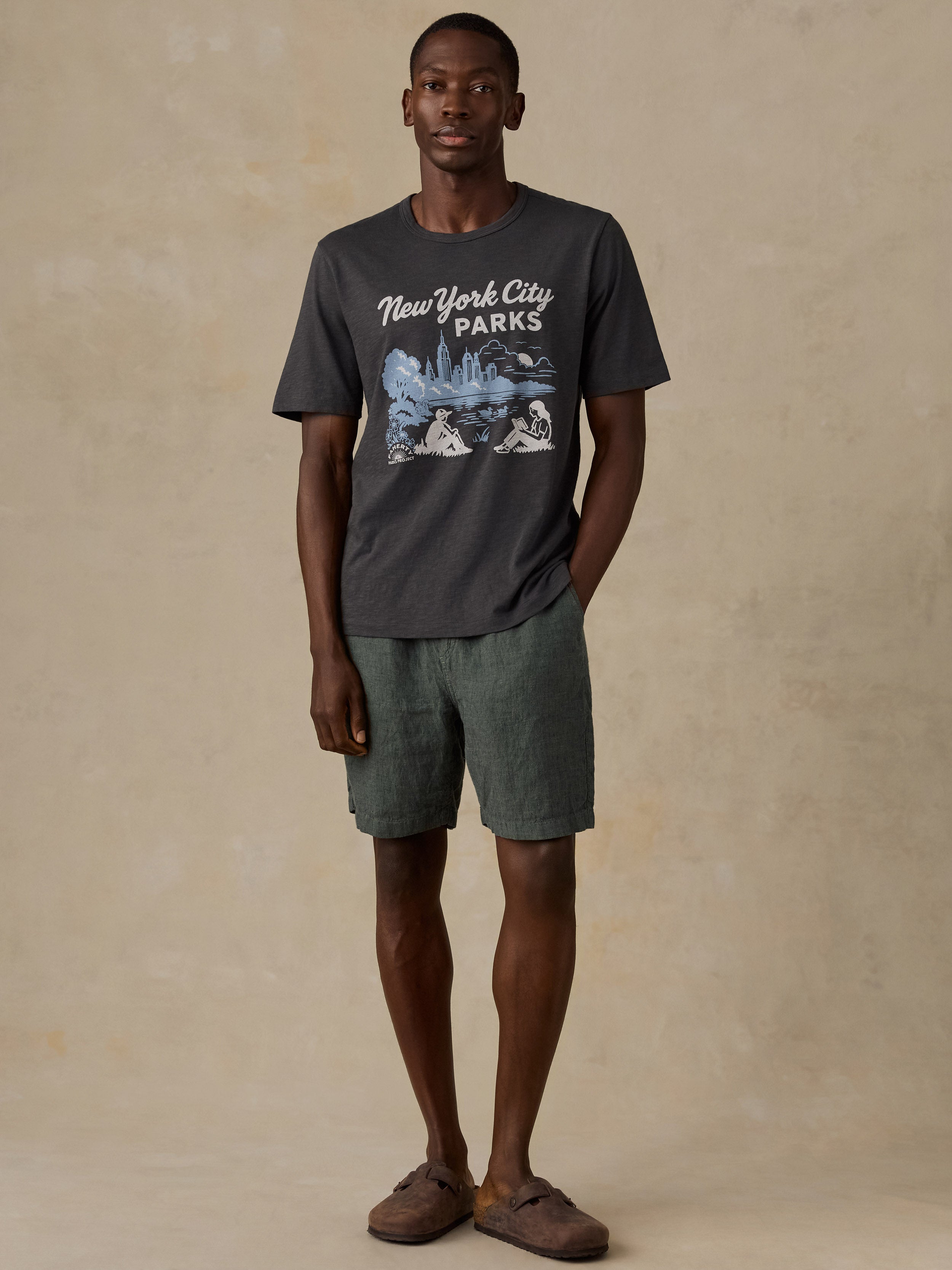 A man wearing a dark gray graphic t-shirt with 'New York City Parks' text and a blue park illustration, paired with olive green shorts and brown slip-on shoes, standing against a textured beige studio backdrop.