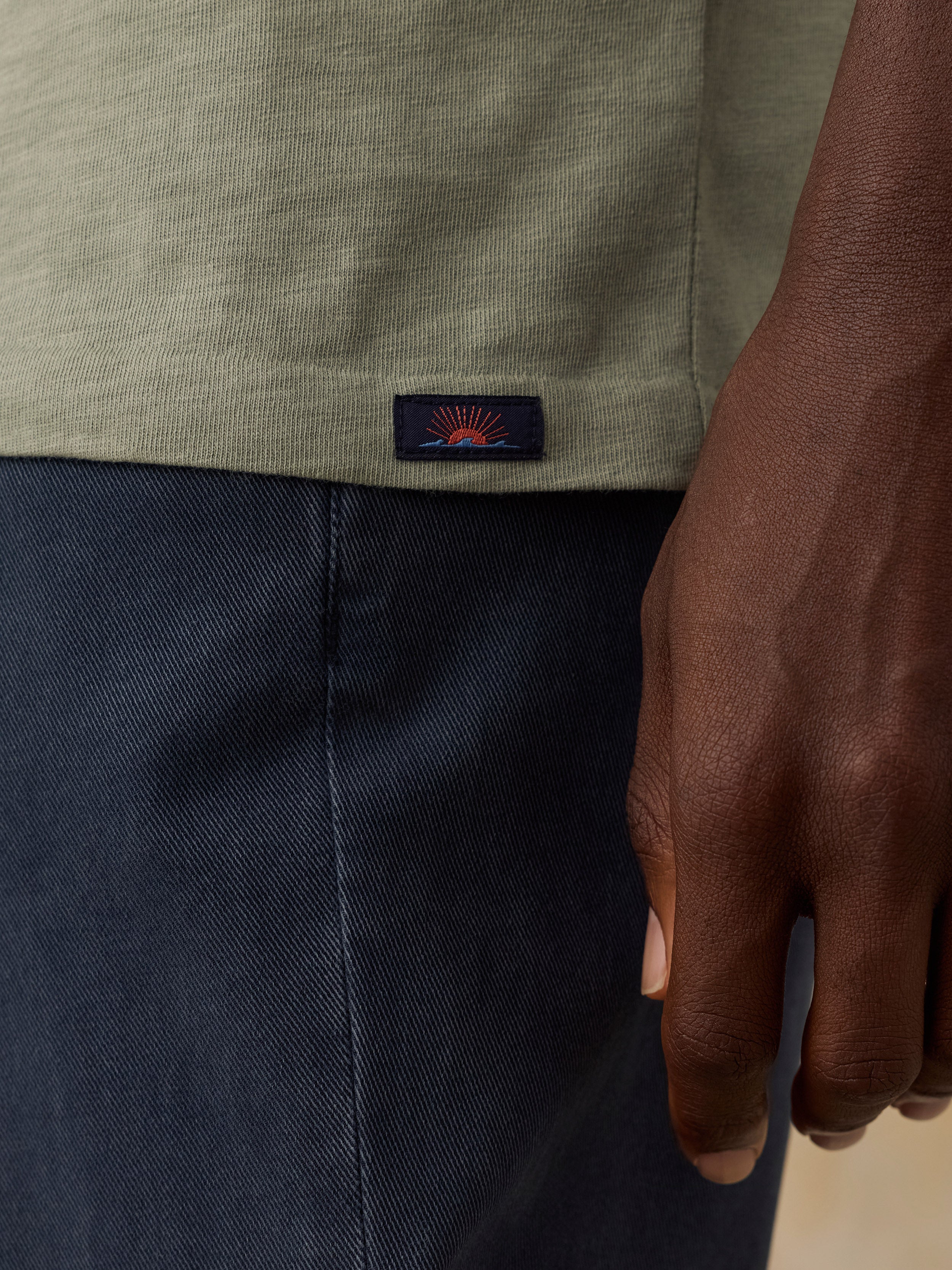 Close-up of a man wearing a light olive green short-sleeve top and navy blue pants, with a small Faherty brand logo patch on the hem.