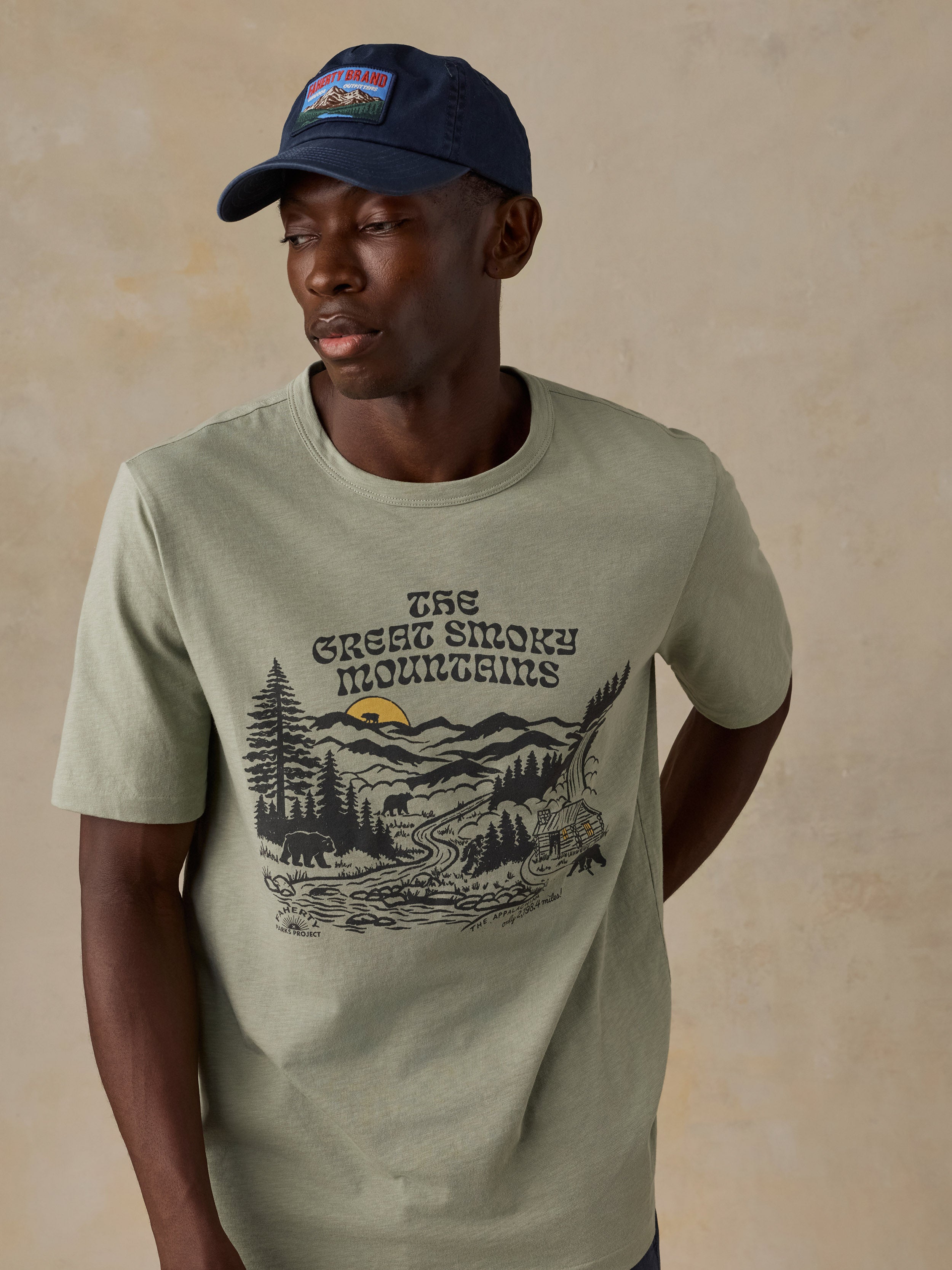A man wearing a light sage green t-shirt with a 'The Great Smoky Mountains' graphic print on the front, paired with a navy blue Faherty cap, set against a textured beige background.