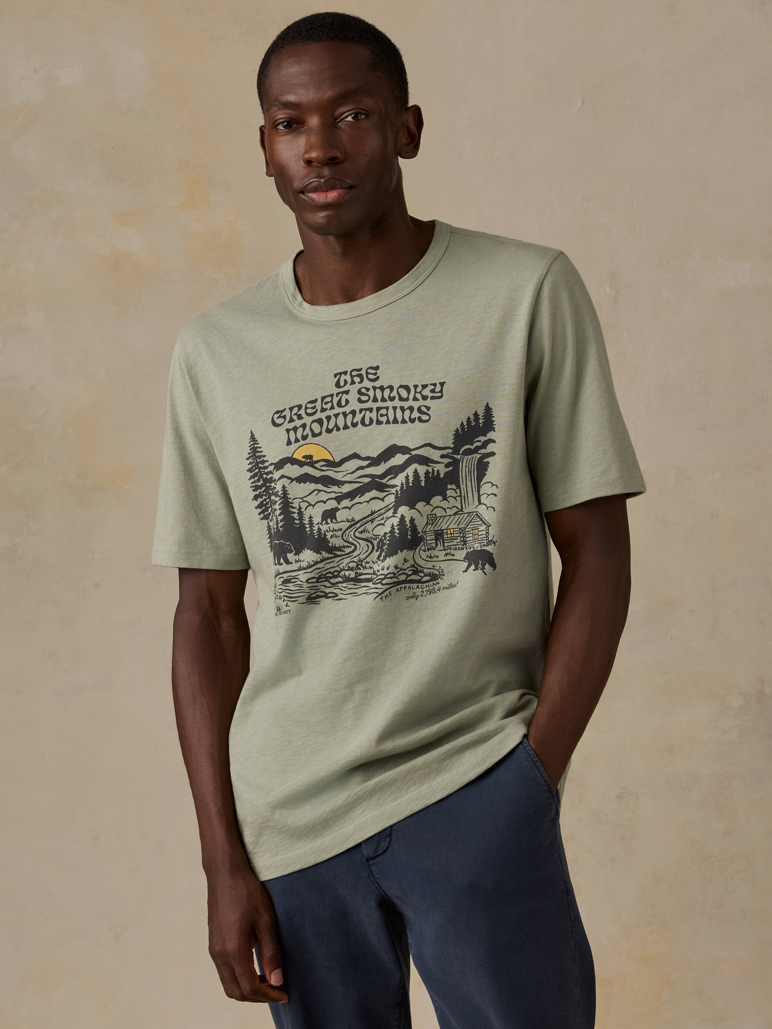 A man wearing a light sage green t-shirt with a 'The Great Smoky Mountains' graphic print on the front, paired with navy blue pants, standing against a textured beige background.