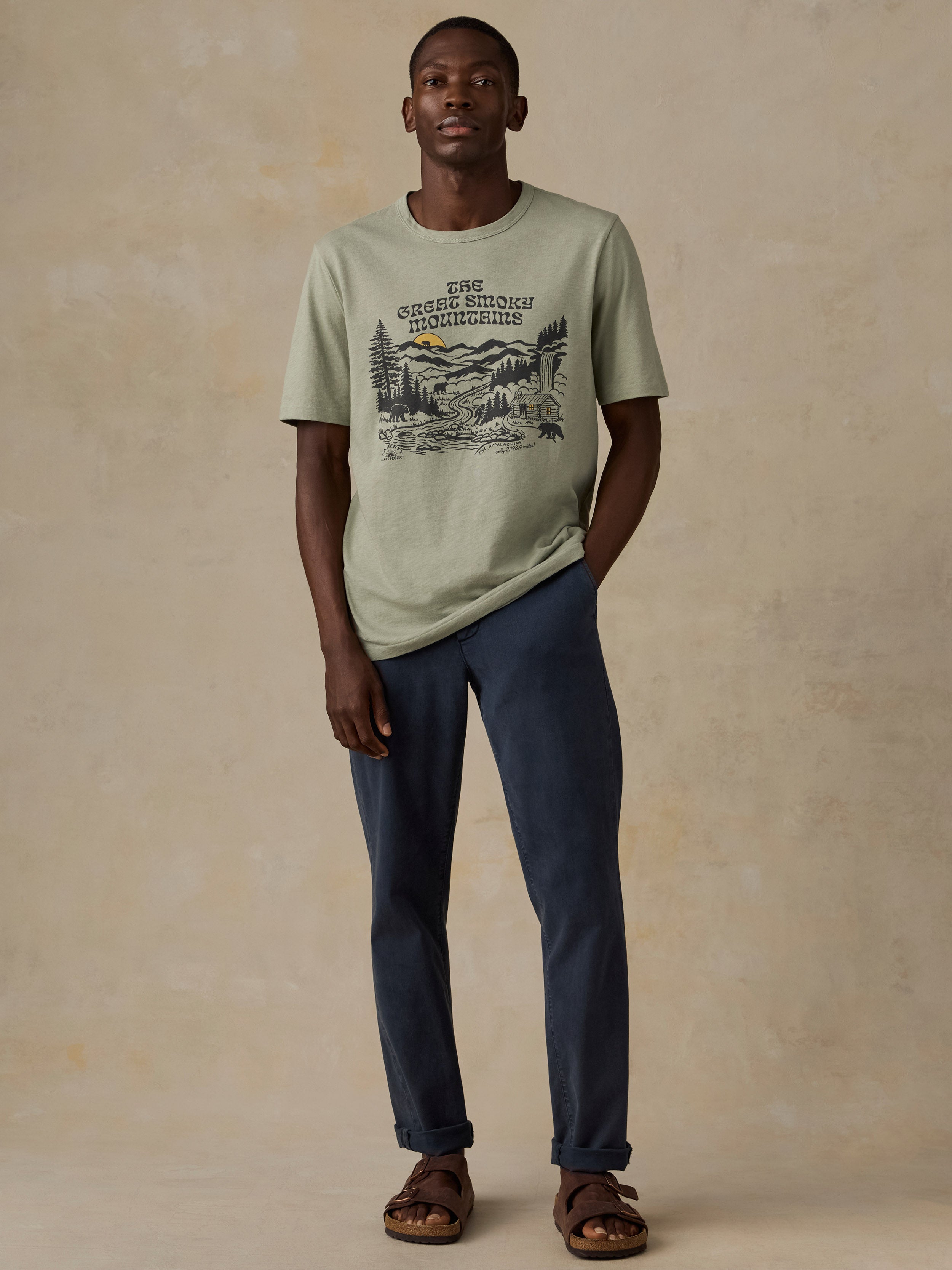 A man modeling a sage green graphic t-shirt with a mountain print and text, paired with navy blue pants and brown sandals, standing against a textured beige studio backdrop.