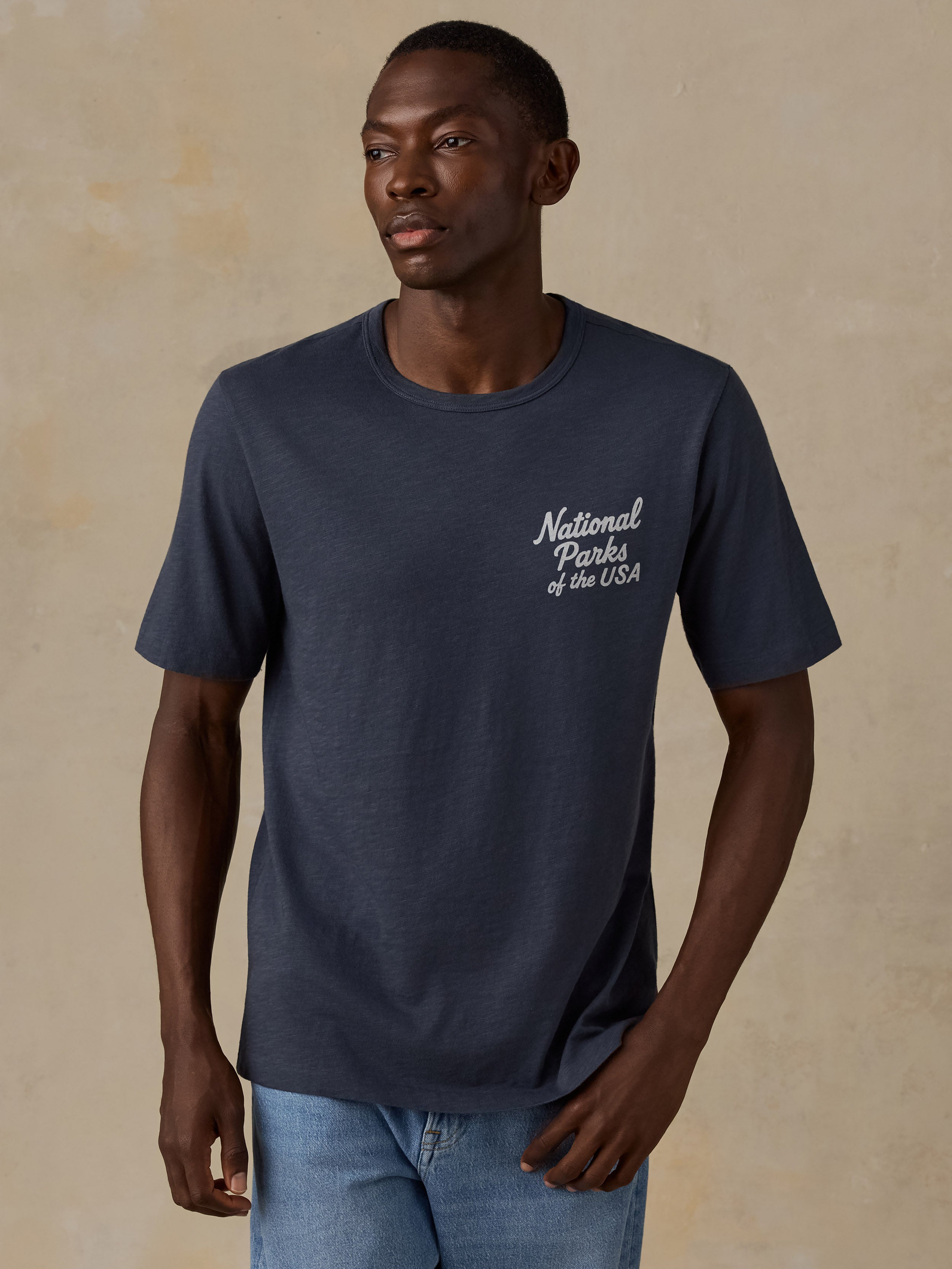 A man wearing a navy blue crewneck t-shirt with 'National Parks of the USA' printed in white script on the left chest, paired with light blue jeans, set against a textured beige background.