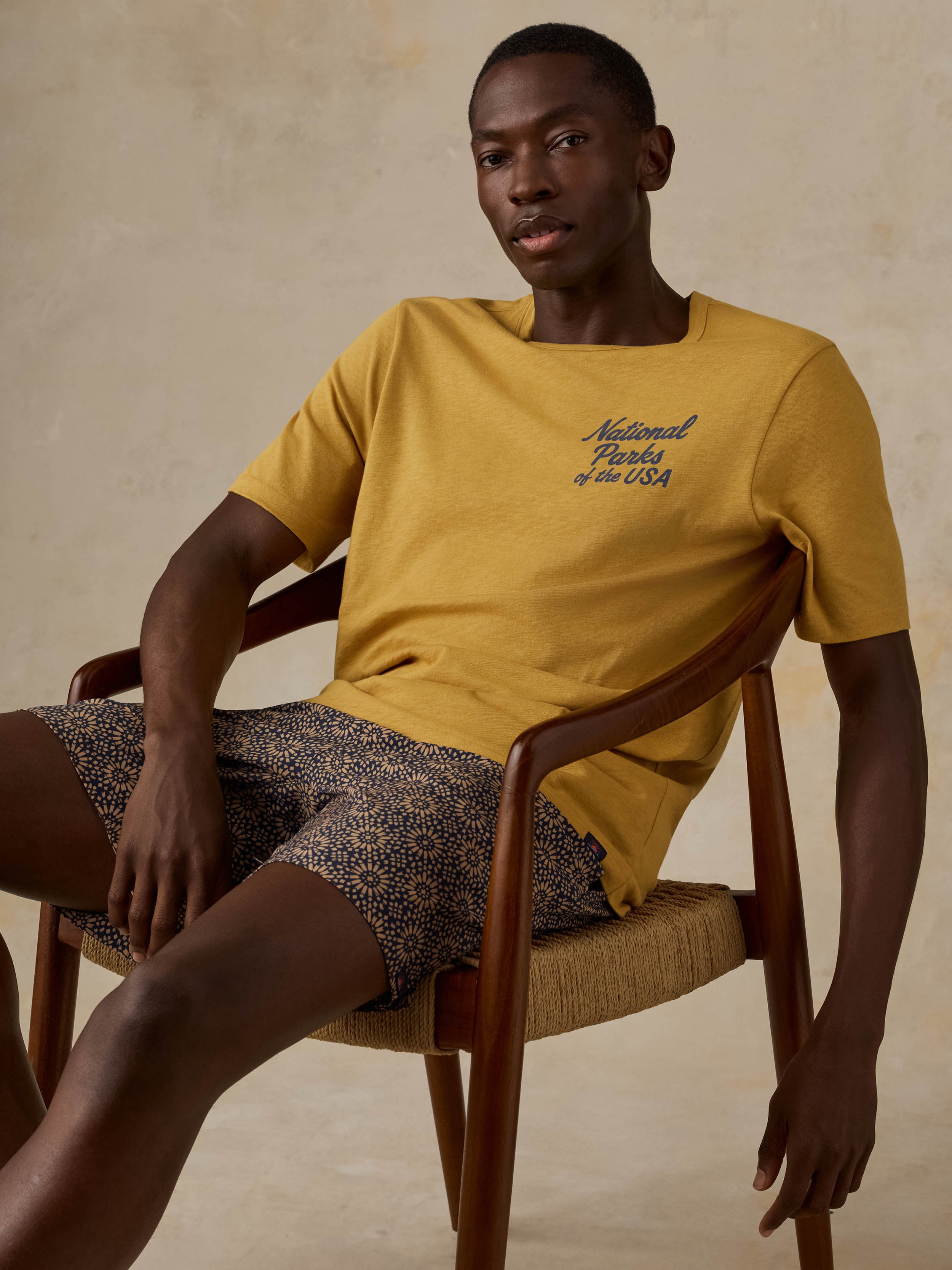 A man sitting on a wooden chair wearing a yellow National Parks tee with printed shorts against a beige background