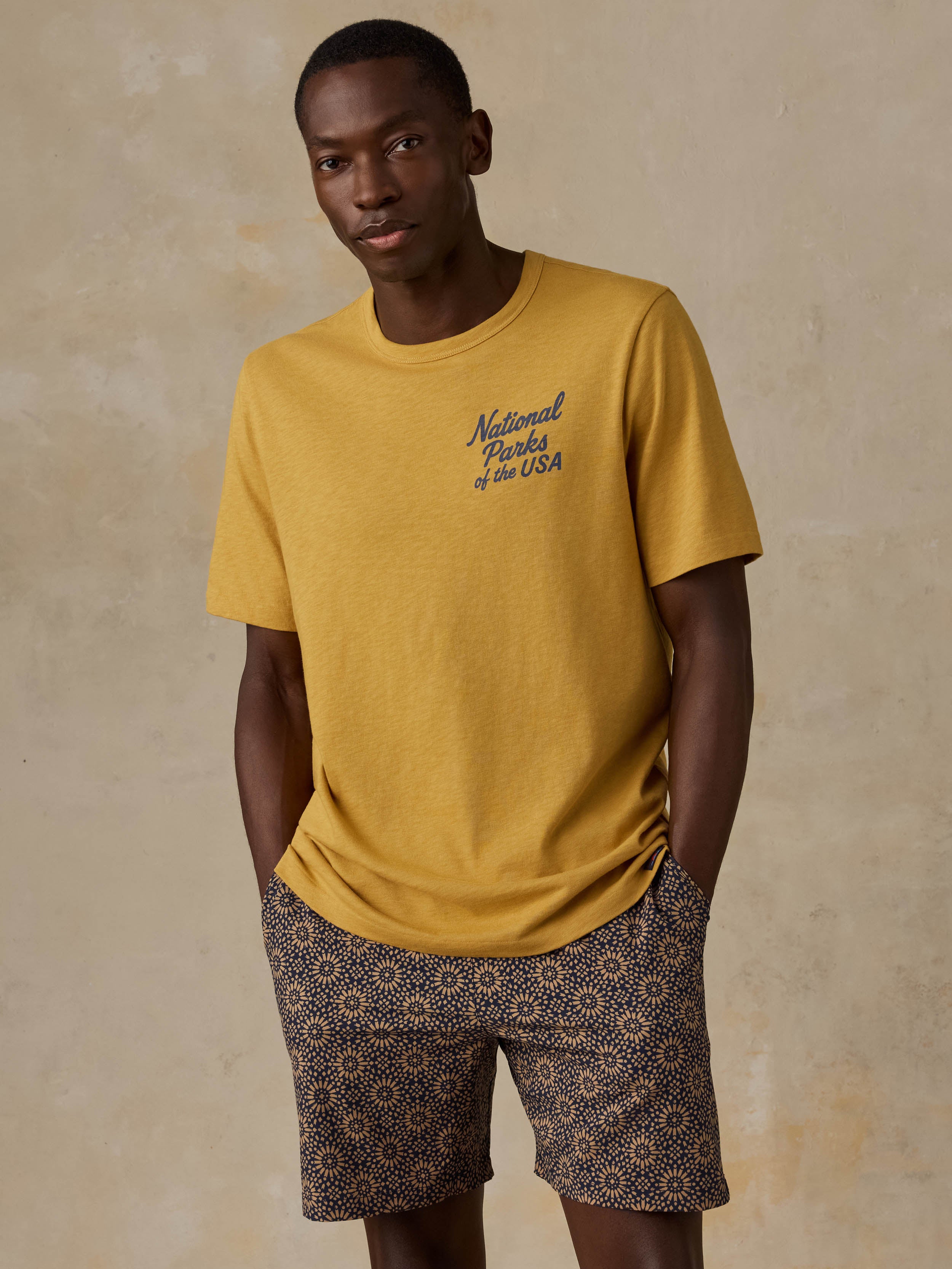 A man wearing a yellow National Parks tee with printed shorts against a beige background