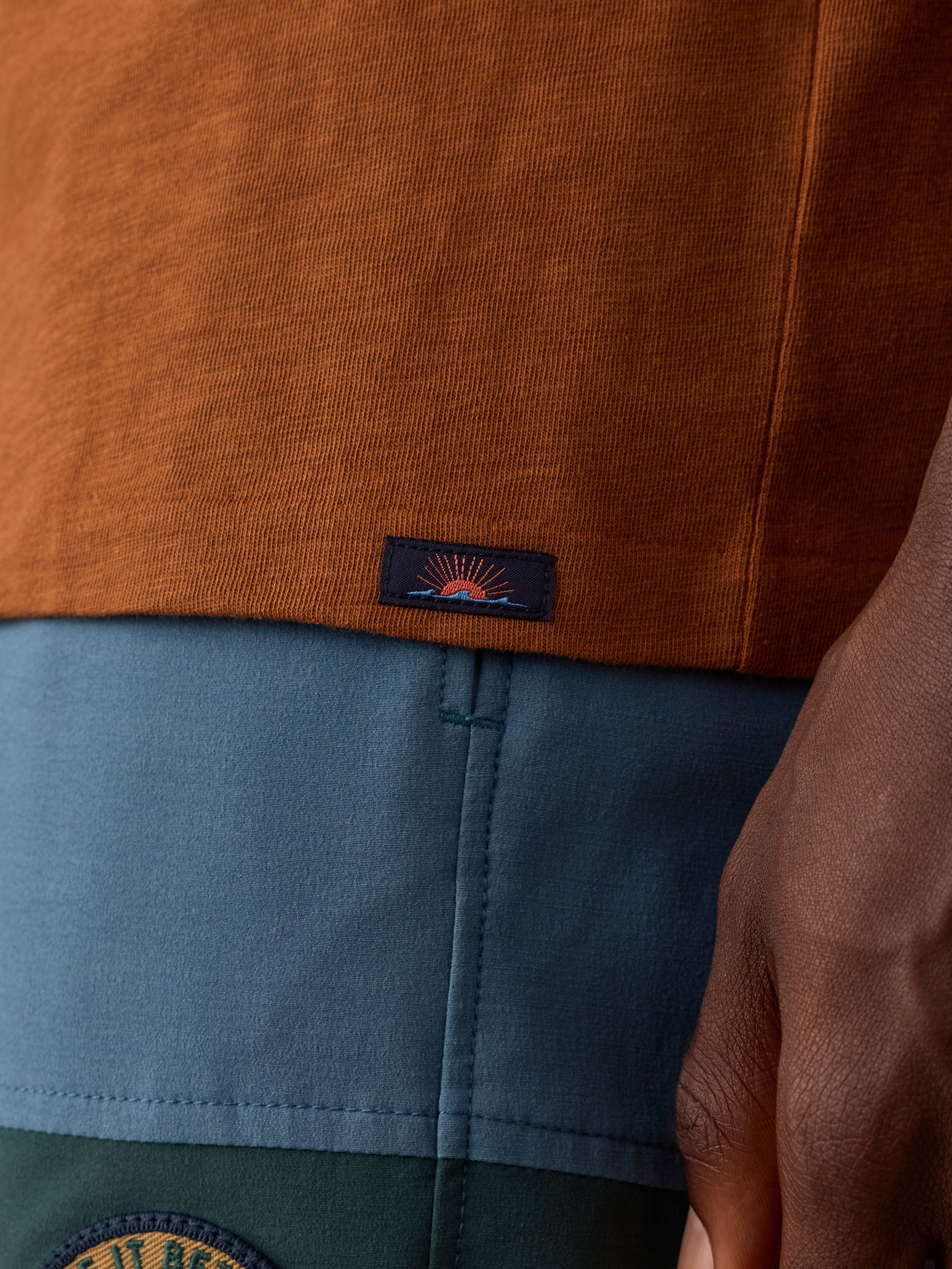 Close-up of a man wearing a rust-orange knit top layered over a blue and green color-blocked bottom, with a small Faherty brand logo patch near the hem.