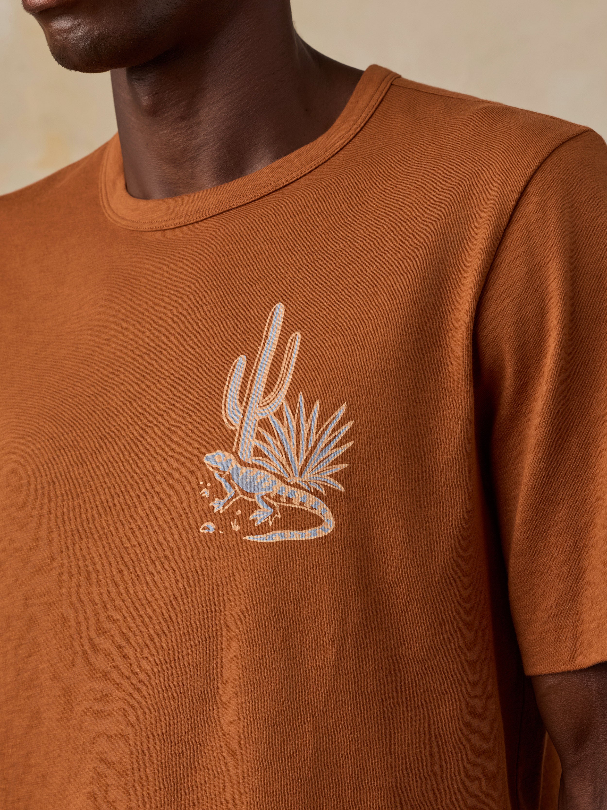 A close-up of a man wearing a rust-orange crewneck t-shirt with a light beige desert graphic featuring a lizard and cactus on the chest.