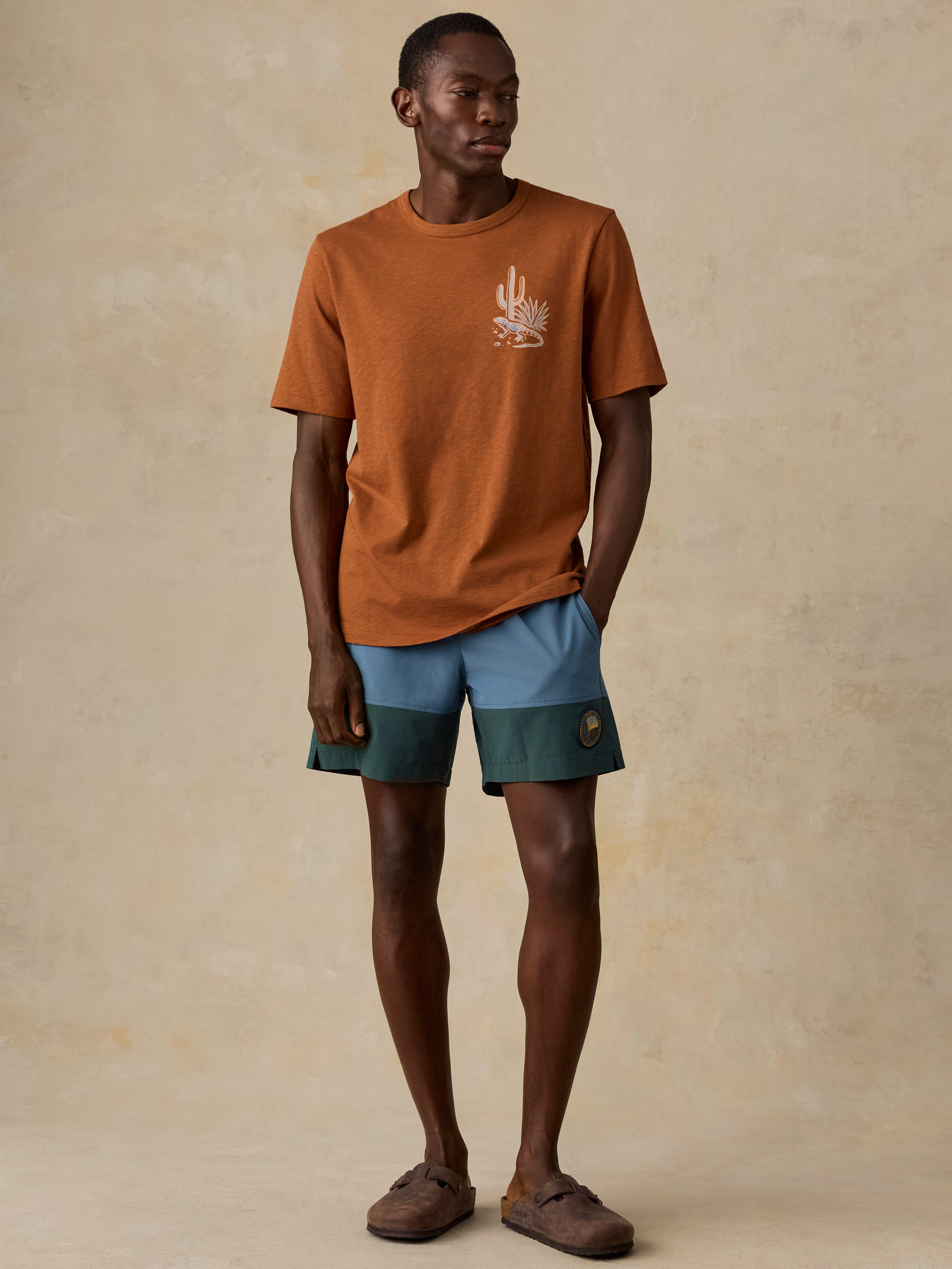A man modeling a rust-orange crewneck t-shirt with a small white graphic on the left chest, paired with color-blocked blue and green shorts and brown suede slide sandals, set against a textured beige studio backdrop.
