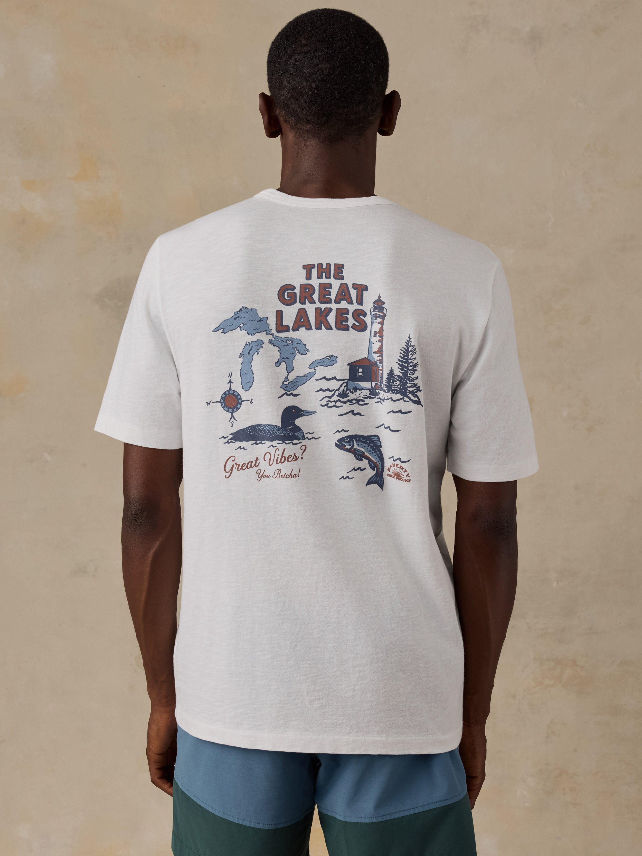 A man viewed from behind wearing a white t-shirt with a large back graphic that reads 'THE GREAT LAKES' and features illustrations of Michigan, a lighthouse, a whale, and a fish. He is standing against a textured beige background.