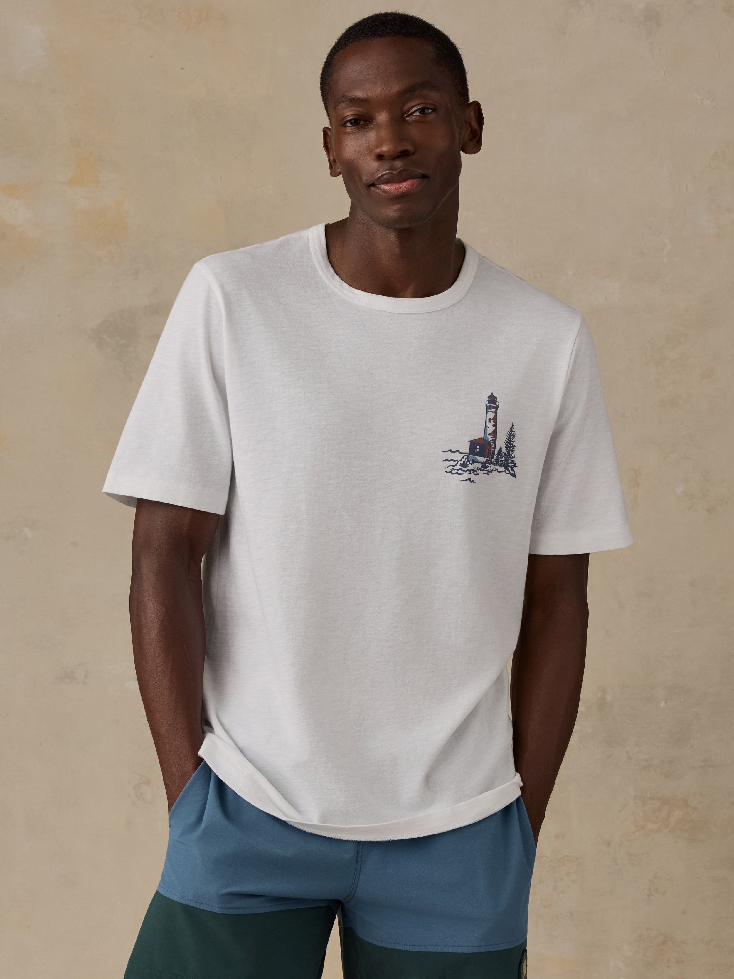 A man wearing a white crew-neck t-shirt with a small dark blue lighthouse graphic on the left chest, paired with blue and green shorts, standing against a textured beige wall.