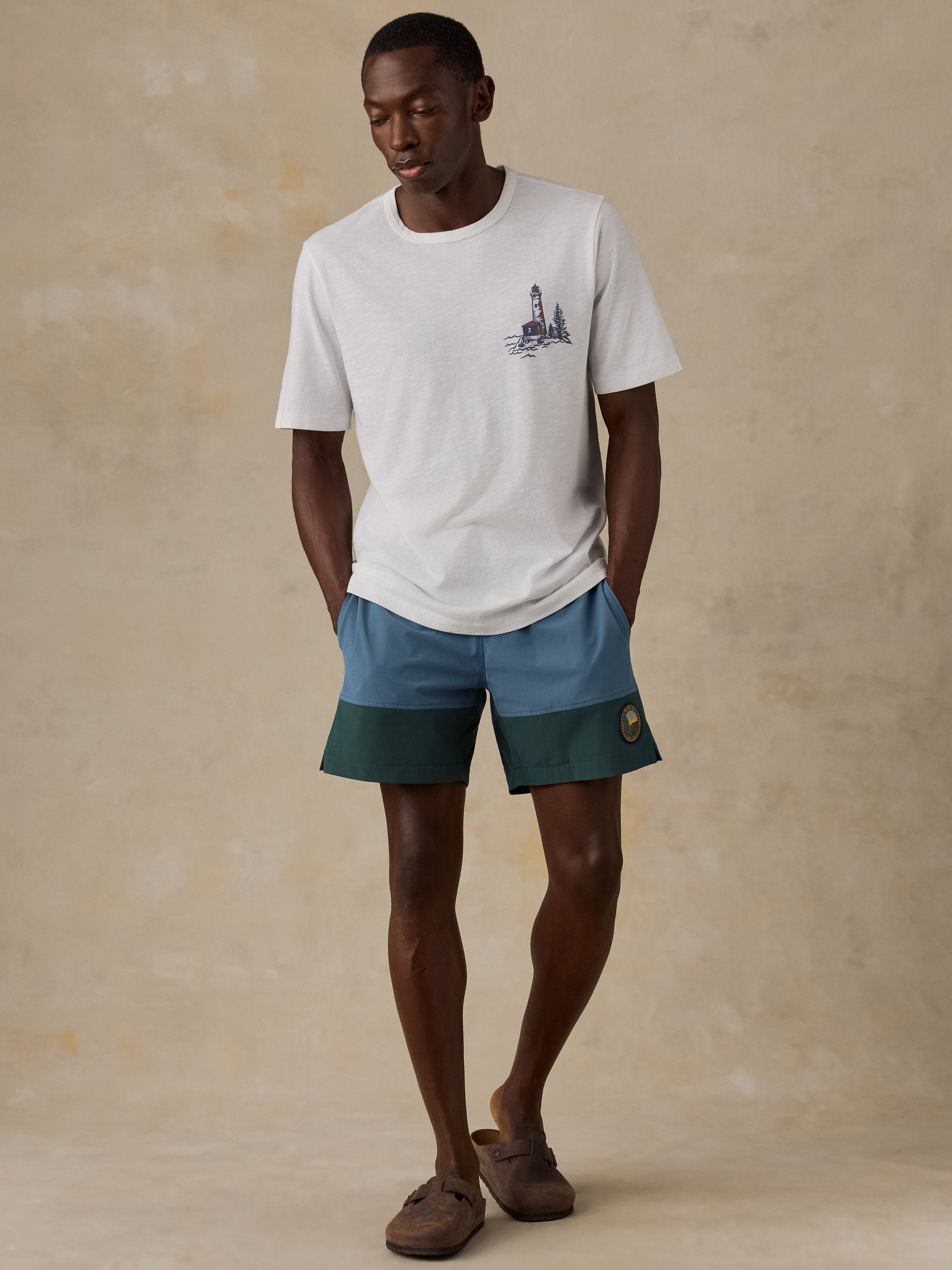 A man modeling a white crewneck t-shirt with a small dark graphic on the left chest, paired with blue and dark green color-blocked shorts and brown slide sandals, set against a textured beige studio backdrop.