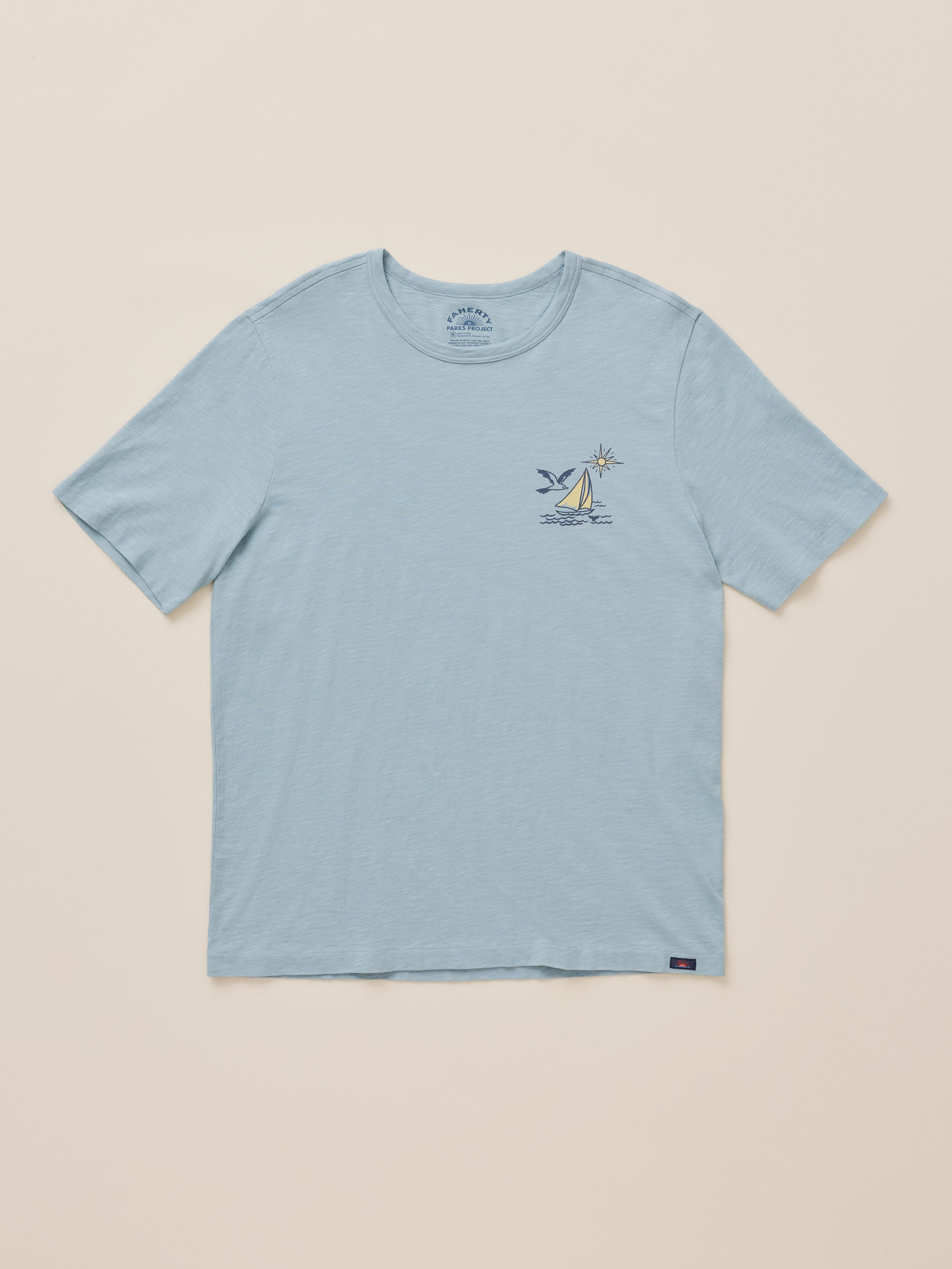 A light blue short-sleeve crewneck t-shirt with a small sailboat graphic on the left chest, displayed flat against a solid beige background.