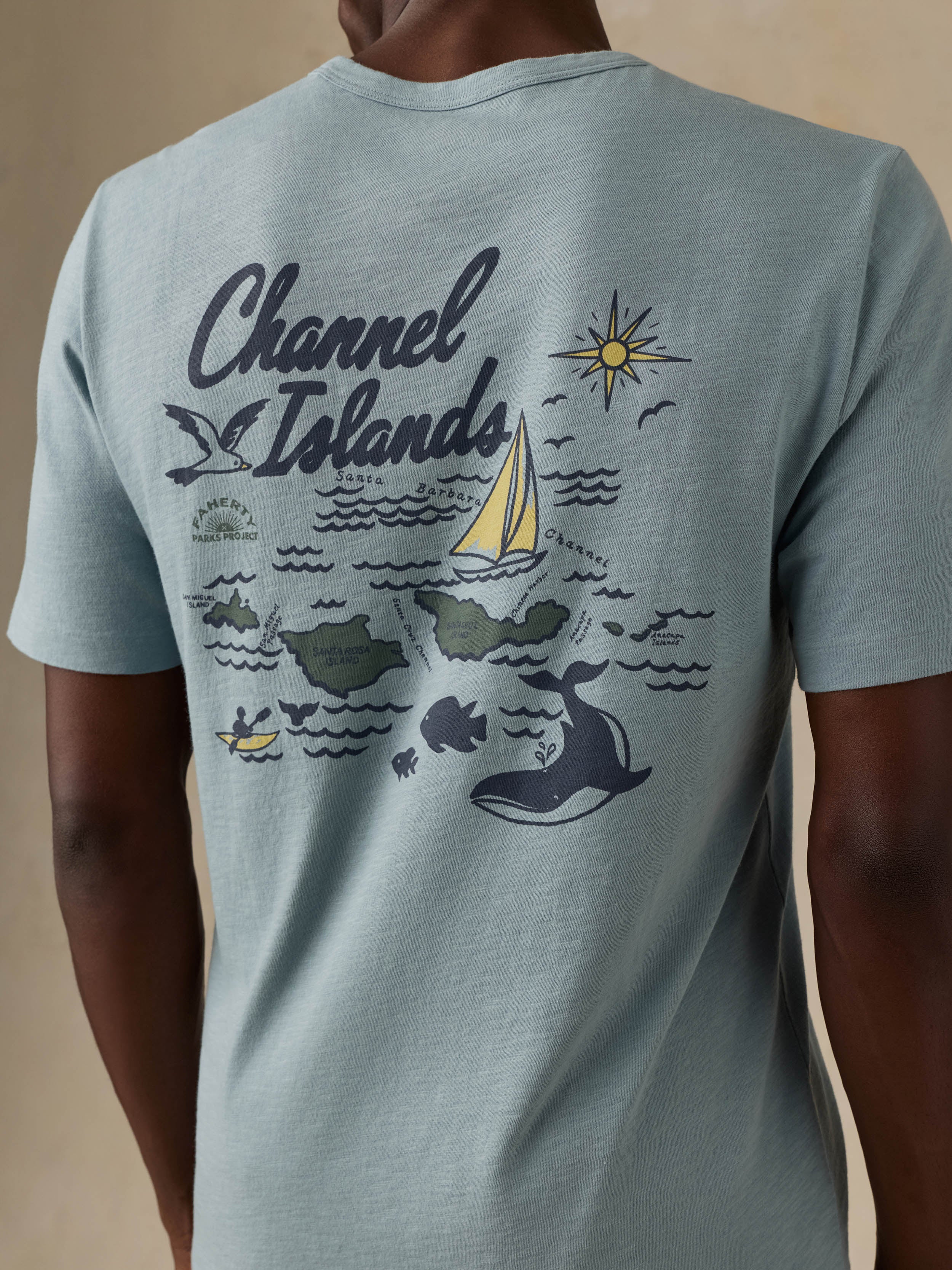 A close-up of a man wearing a light blue t-shirt, showing the back graphic with 'Channel Islands' text and nautical illustrations.