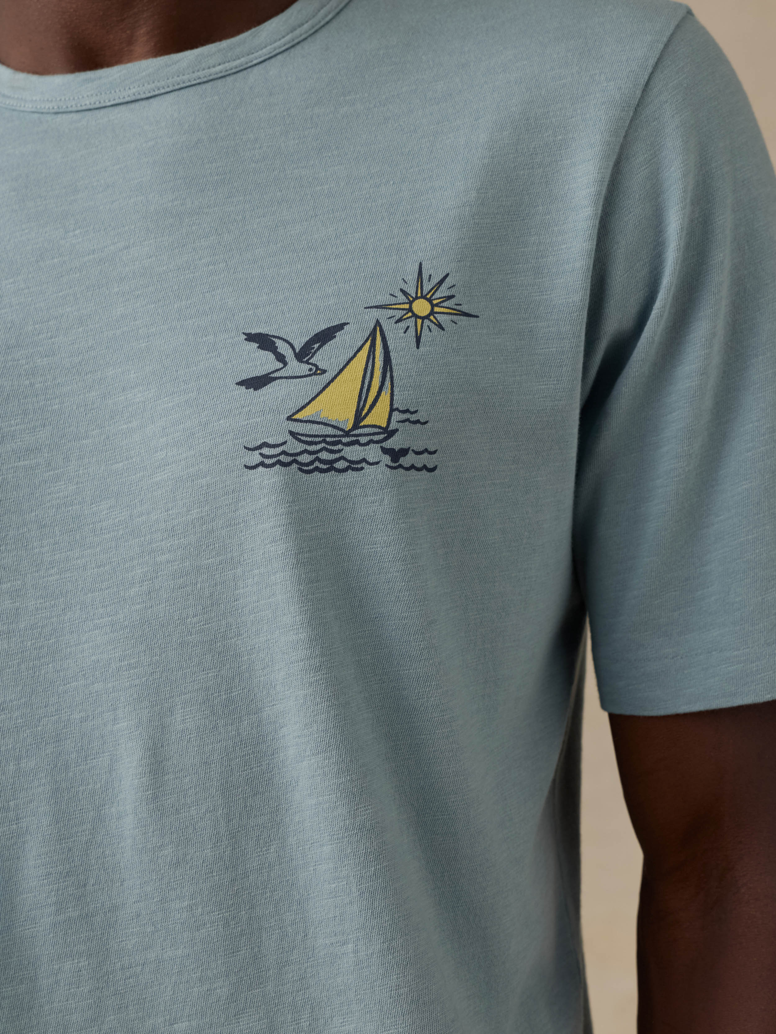 A close-up of a man wearing a light blue-gray short-sleeve t-shirt with a graphic of a yellow sailboat, seagull, and a sun on the chest.