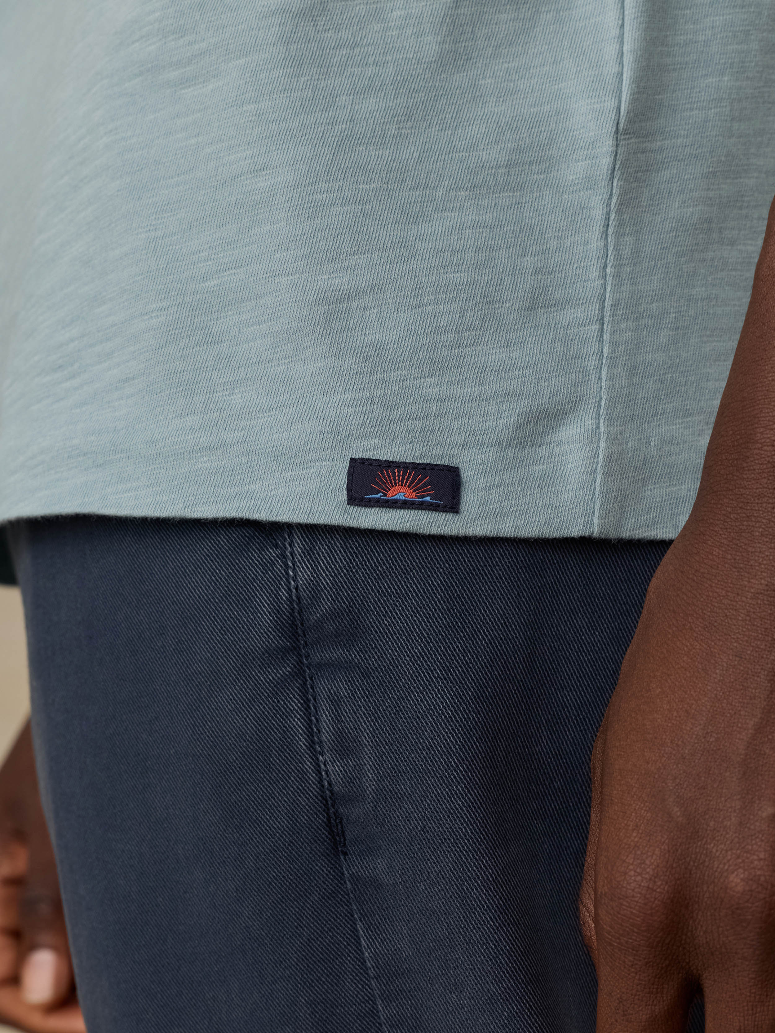 Close-up of a man wearing a light blue t-shirt and navy blue pants, showing a small embroidered Faherty brand patch on the hem.