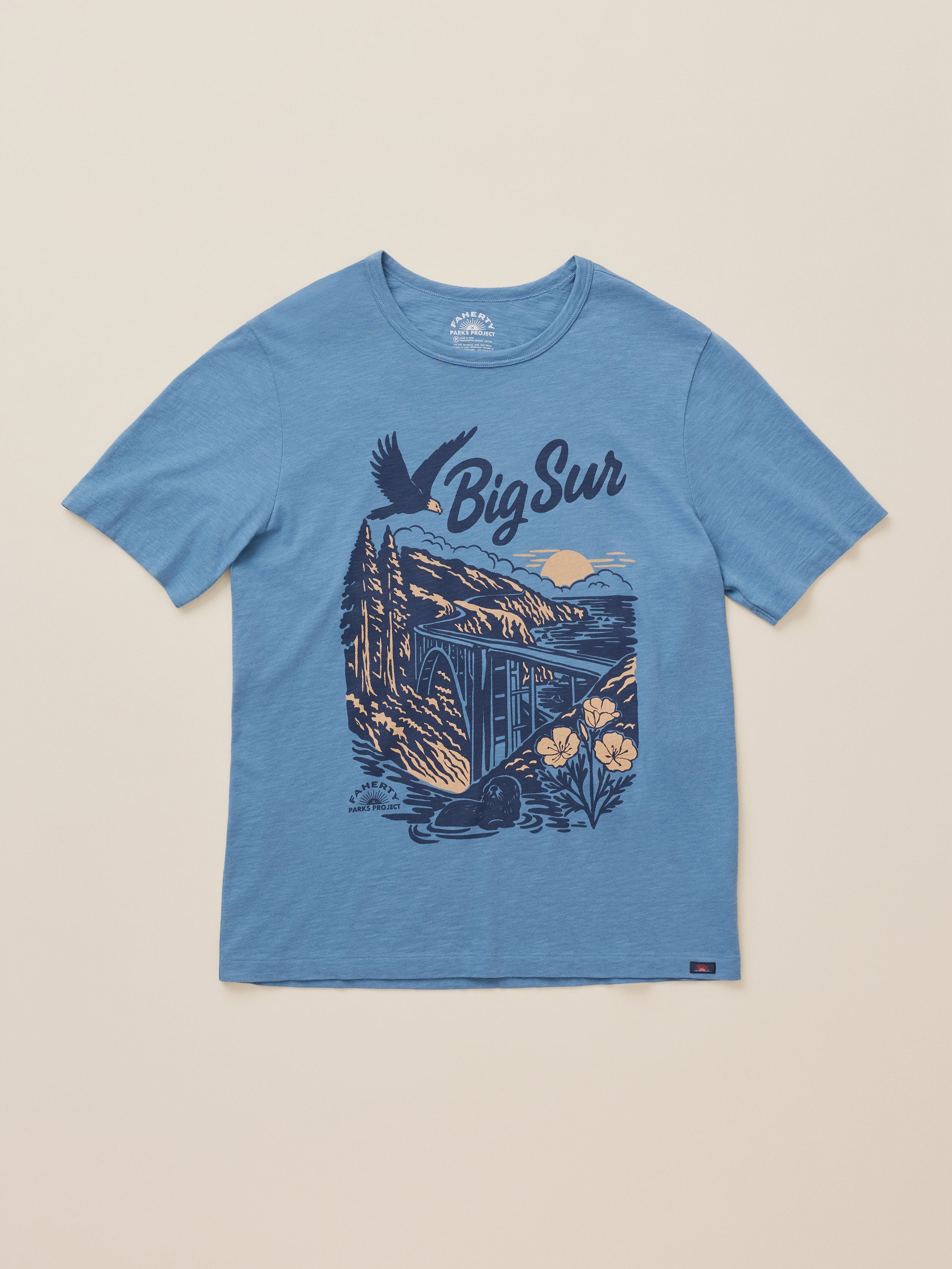 A heather blue short-sleeve t-shirt with a dark blue graphic print on the front featuring the text 'Big Sur' and an illustration of a coastal landscape, displayed flat against a light beige background.