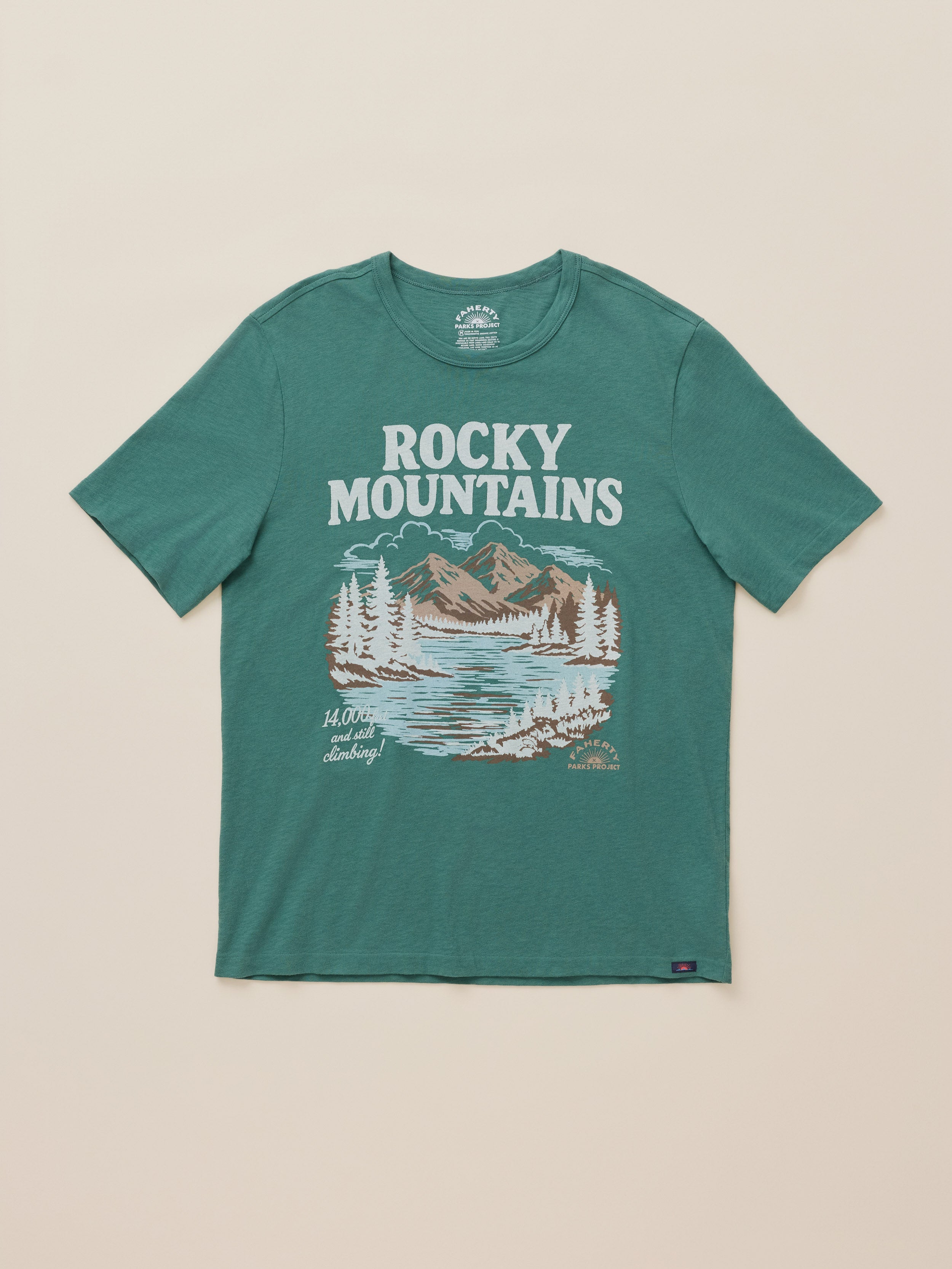 A teal green short-sleeve t-shirt with a 'Rocky Mountains' graphic on the front, featuring a mountain and lake illustration in white, beige, and brown, laid flat against a light beige background.