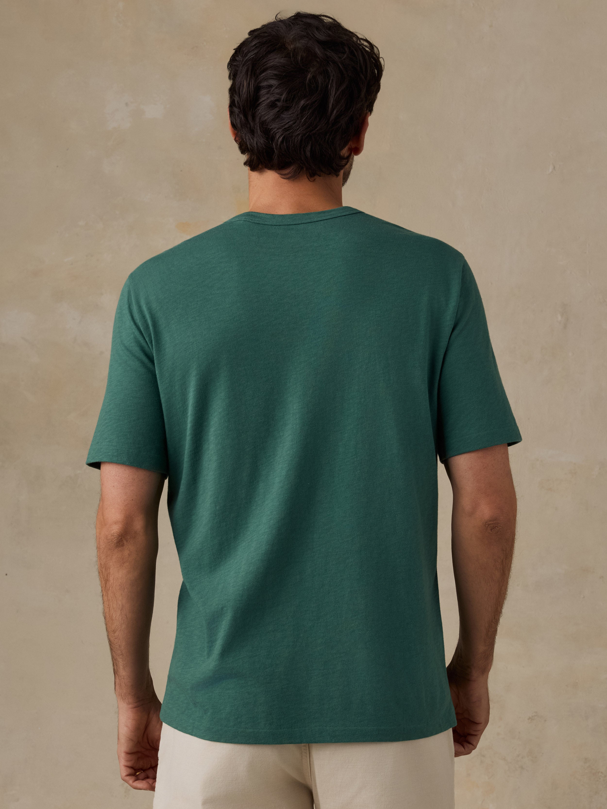 A man viewed from behind wearing a dark green short-sleeve t-shirt and light beige shorts, standing against a textured beige wall.