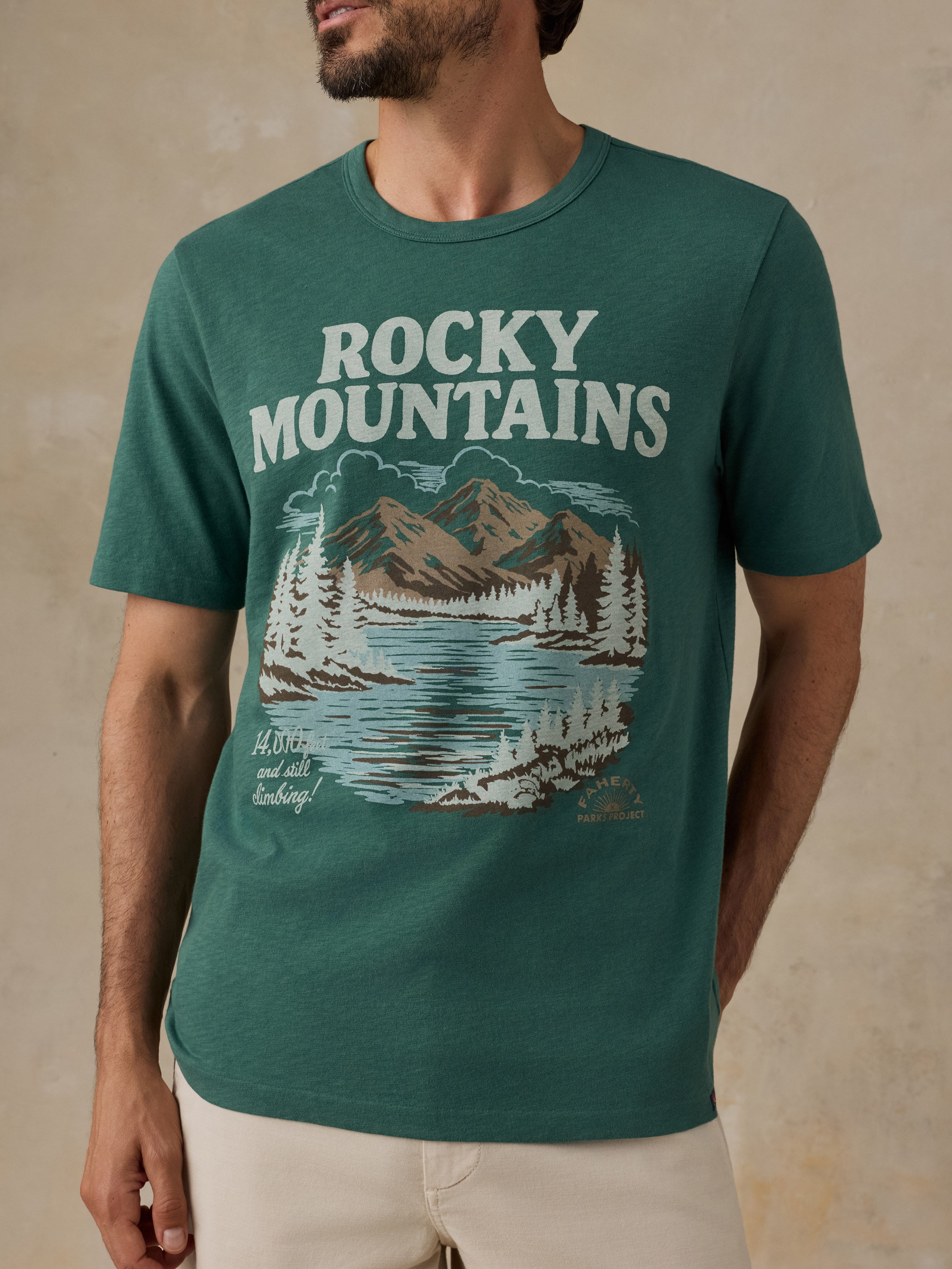 A man wearing a dark teal green t-shirt with a 'Rocky Mountains' graphic print on the front, paired with beige shorts, set against a textured beige background.