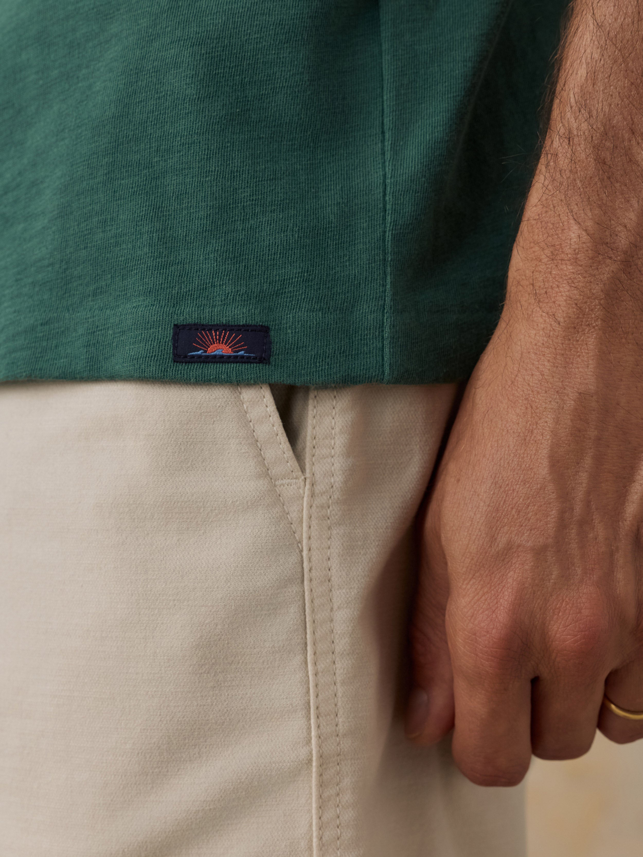 Close-up of a man wearing a dark green short-sleeve shirt and light beige shorts, with a small Faherty brand patch on the shirt near the hem and a hand resting by the pocket.