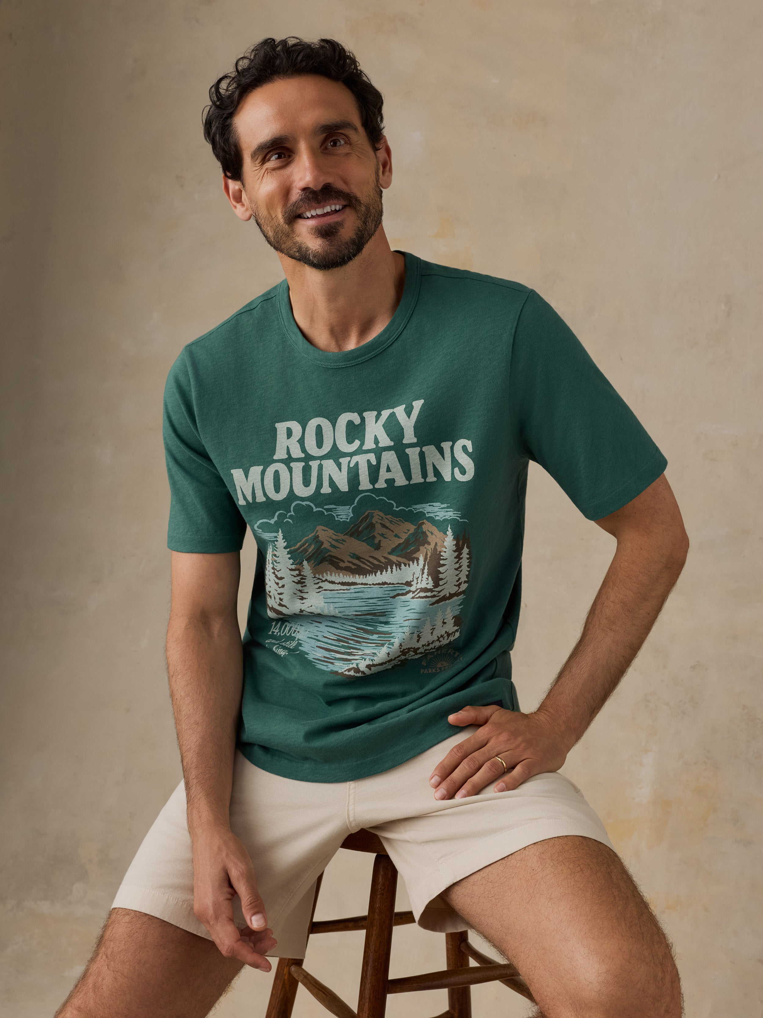 A man wearing a dark green graphic t-shirt with 'Rocky Mountains' and a mountain landscape print, paired with beige shorts, sitting on a wooden stool against a textured beige wall.