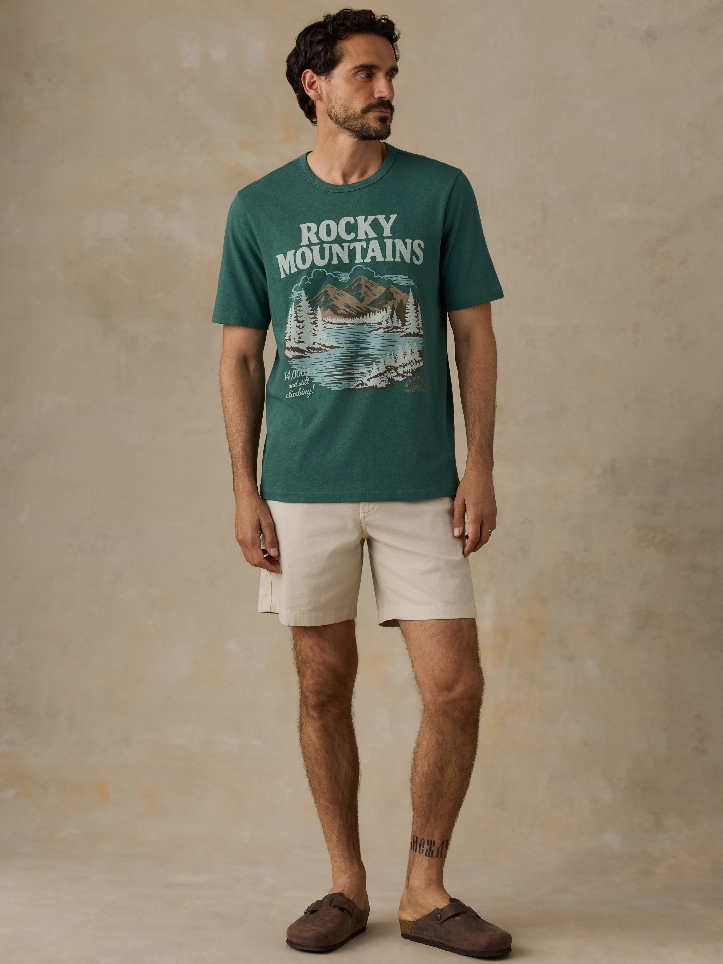 A man wearing a dark green graphic t-shirt with 'Rocky Mountains' and a mountain/sailboat print, paired with beige shorts and brown slip-on shoes, standing against a textured beige studio backdrop.