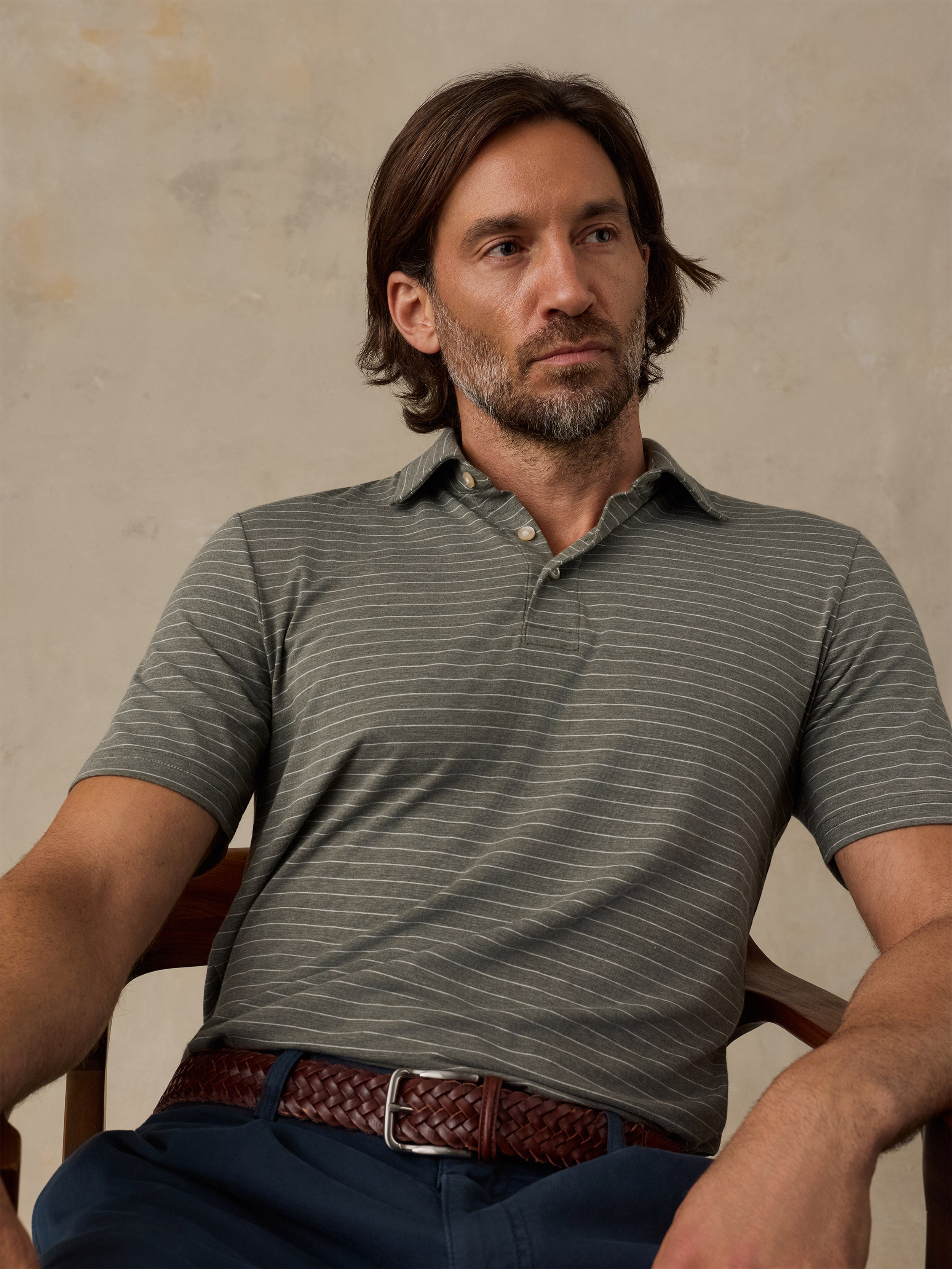 A man wearing a short-sleeve polo shirt with thin horizontal gray stripes on an olive green base, paired with a brown braided belt and navy pants, seated in a wooden chair against a textured beige wall.