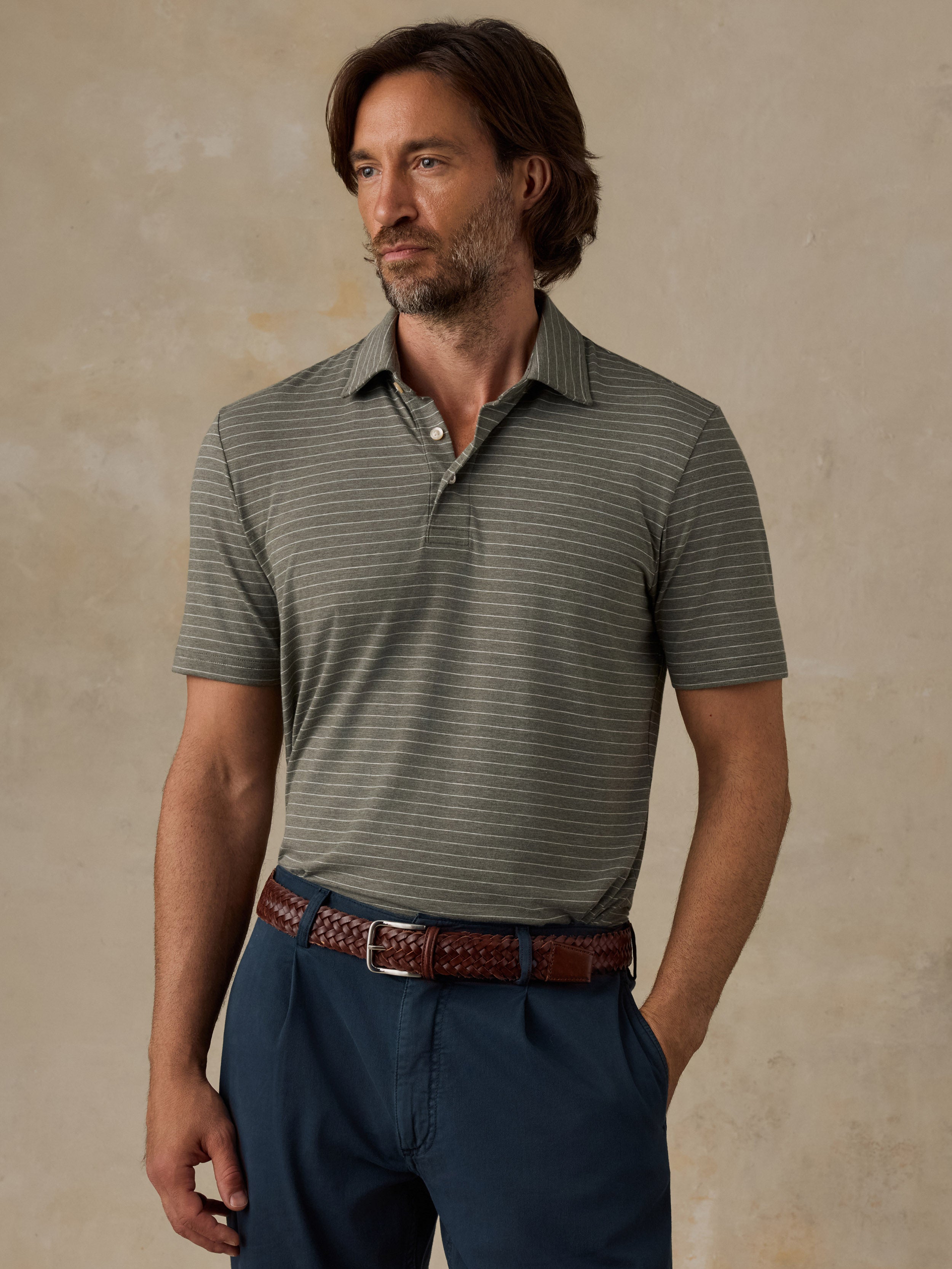 A man wearing a short-sleeve polo shirt with fine horizontal stripes in olive green and dark gray, paired with navy blue trousers and a brown woven belt, standing against a textured beige wall.