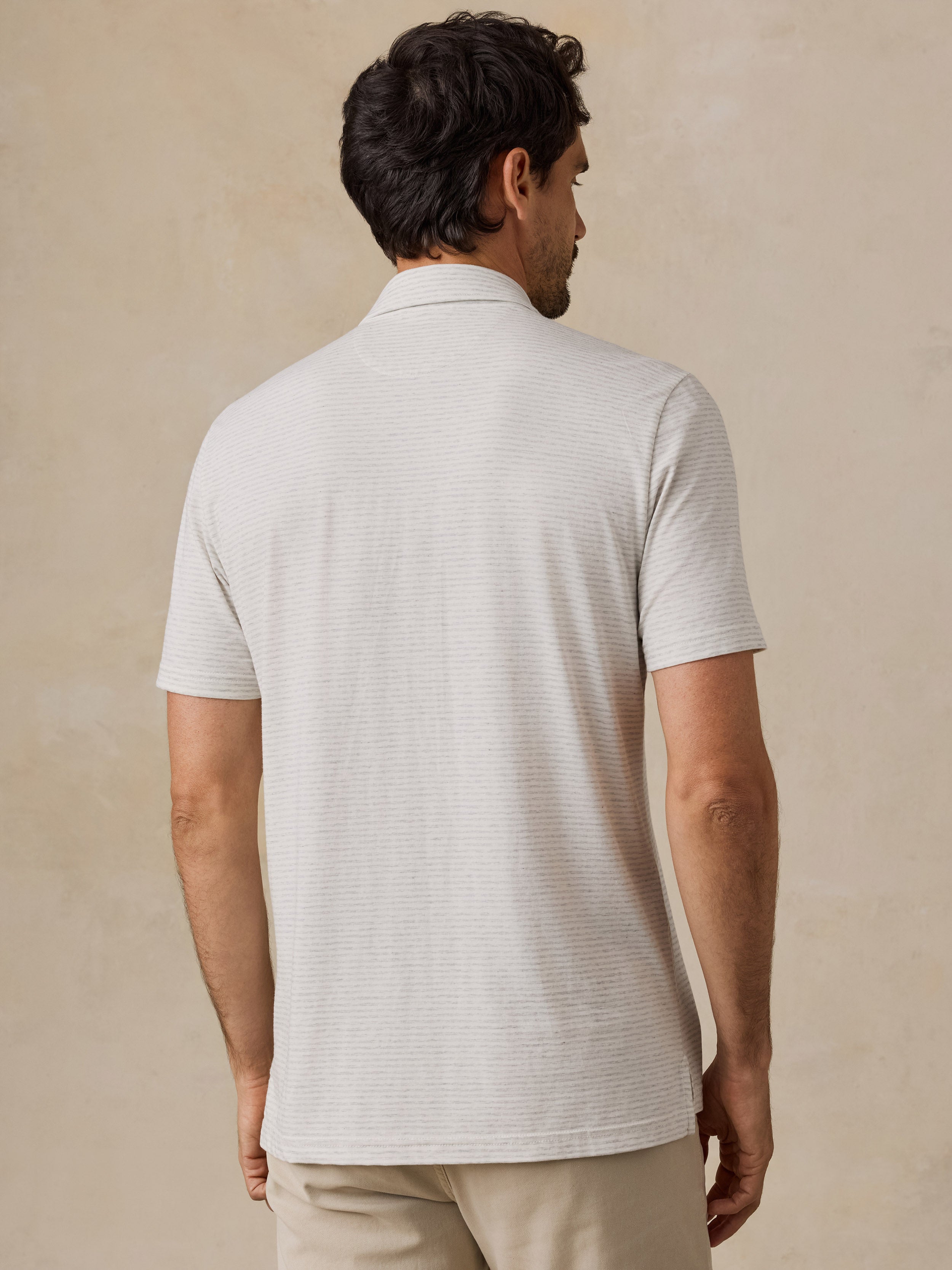 A man viewed from the back wearing a short-sleeve polo shirt in a light cream or off-white color, paired with khaki pants, against a textured beige studio background.