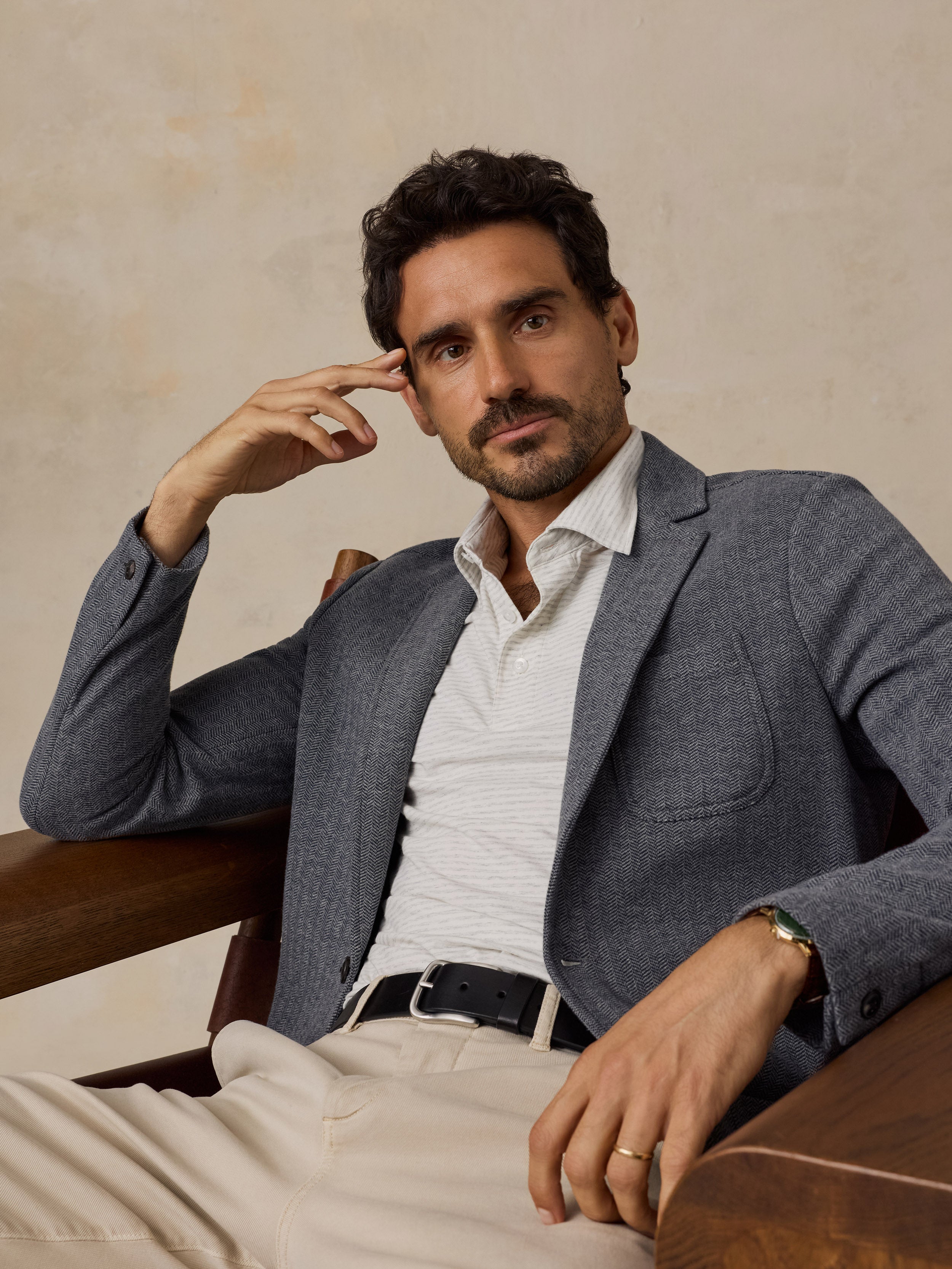 A man wearing a textured gray blazer, white striped polo shirt, and beige trousers, sitting in a wooden armchair against a textured beige wall.