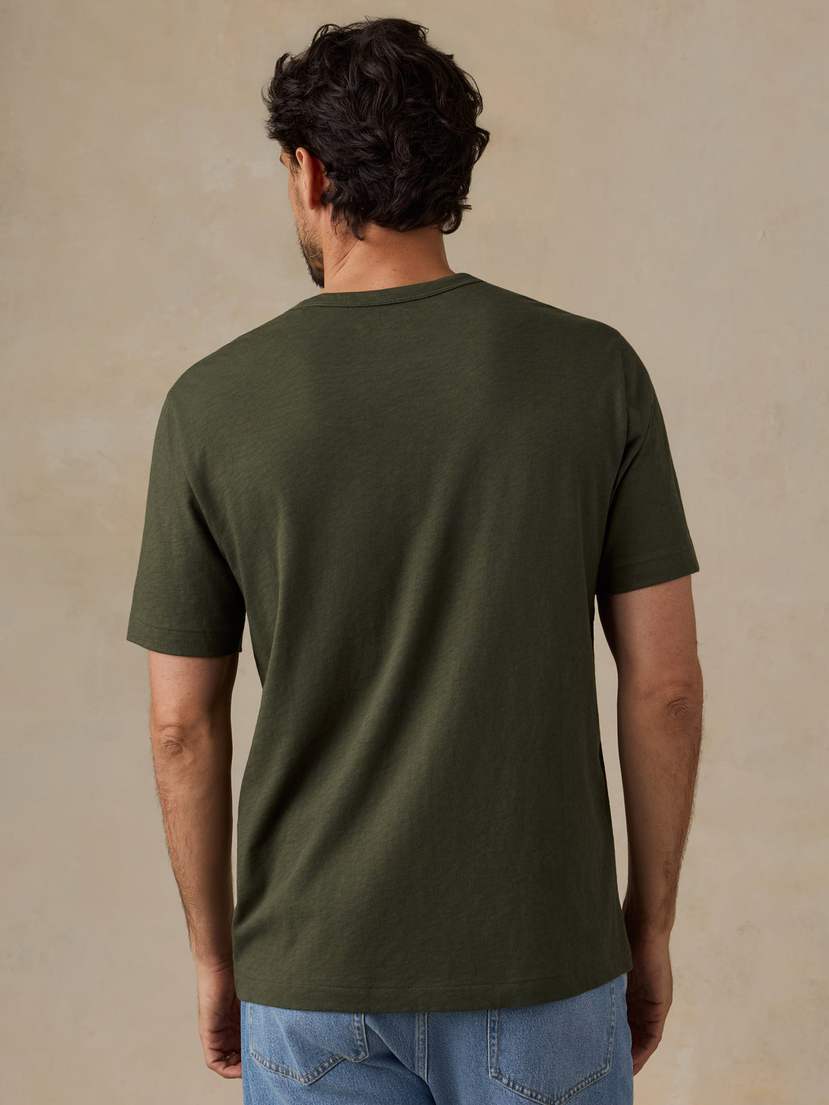 A man viewed from behind wearing a dark olive green short-sleeve t-shirt and light blue jeans, standing against a textured beige studio background., image 3 of 6