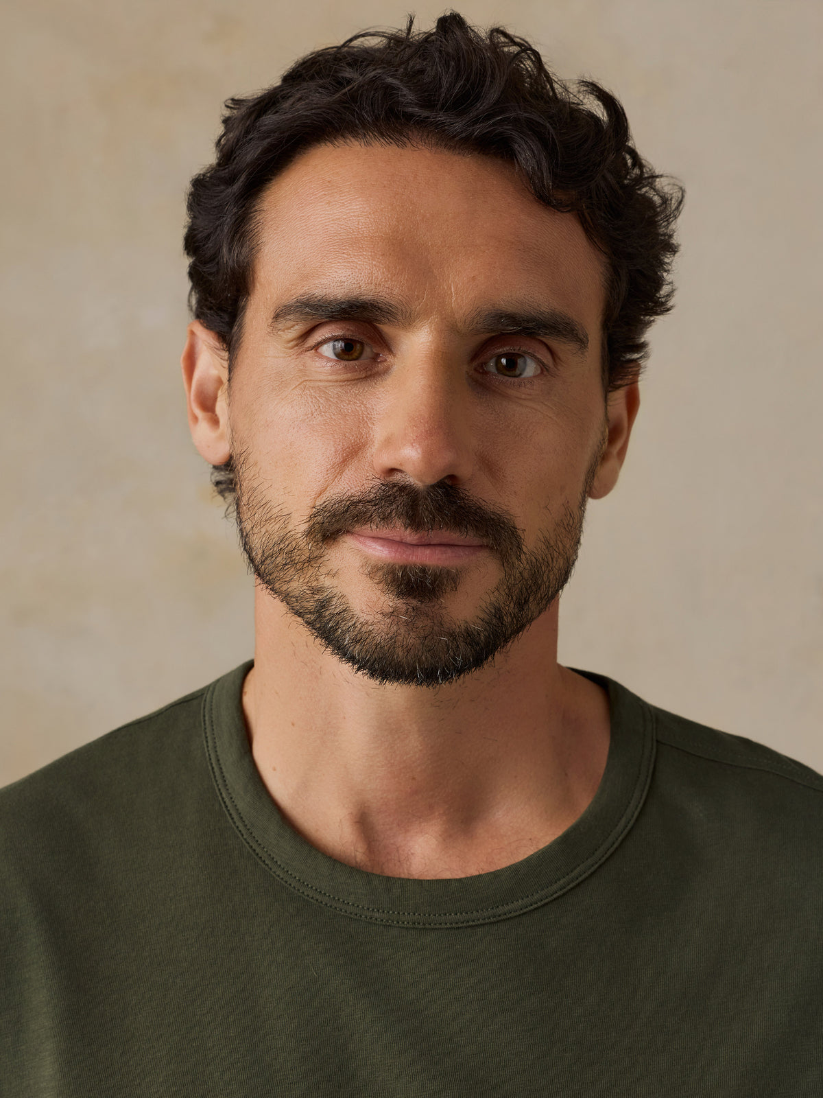 A close-up portrait of a man with dark curly hair, brown eyes, and a short beard, wearing an olive green crew-neck t-shirt against a neutral beige background., image 6 of 6